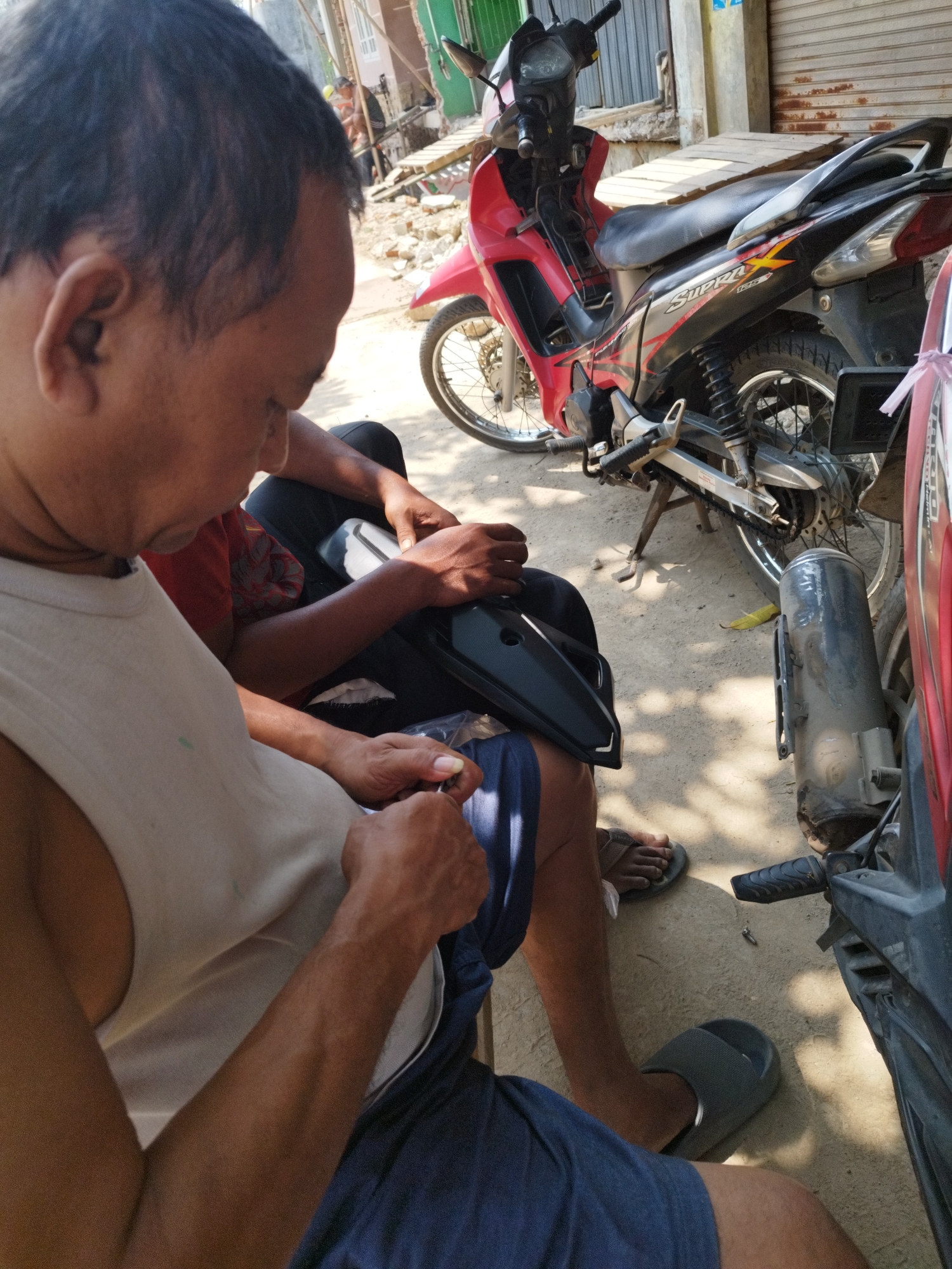 This image depicts an outdoor scene in Karawang, Indonesia, likely on a street or yard. In the foreground, a man with dark hair is shown from the chest up, wearing a white tank top and blue shorts. He is holding a black object, possibly a motorcycle part, and appears to be working on it with his hands. Another person is partially visible behind him, wearing a red shirt. To the right of the man, a red motorcycle is parked, with its exhaust pipe and rear wheel clearly visible. The ground is dirt and sand, with shadows cast from overhead objects, indicating it is daytime. In the background, there are elements of buildings and construction, suggesting an urban or semi-urban environment.