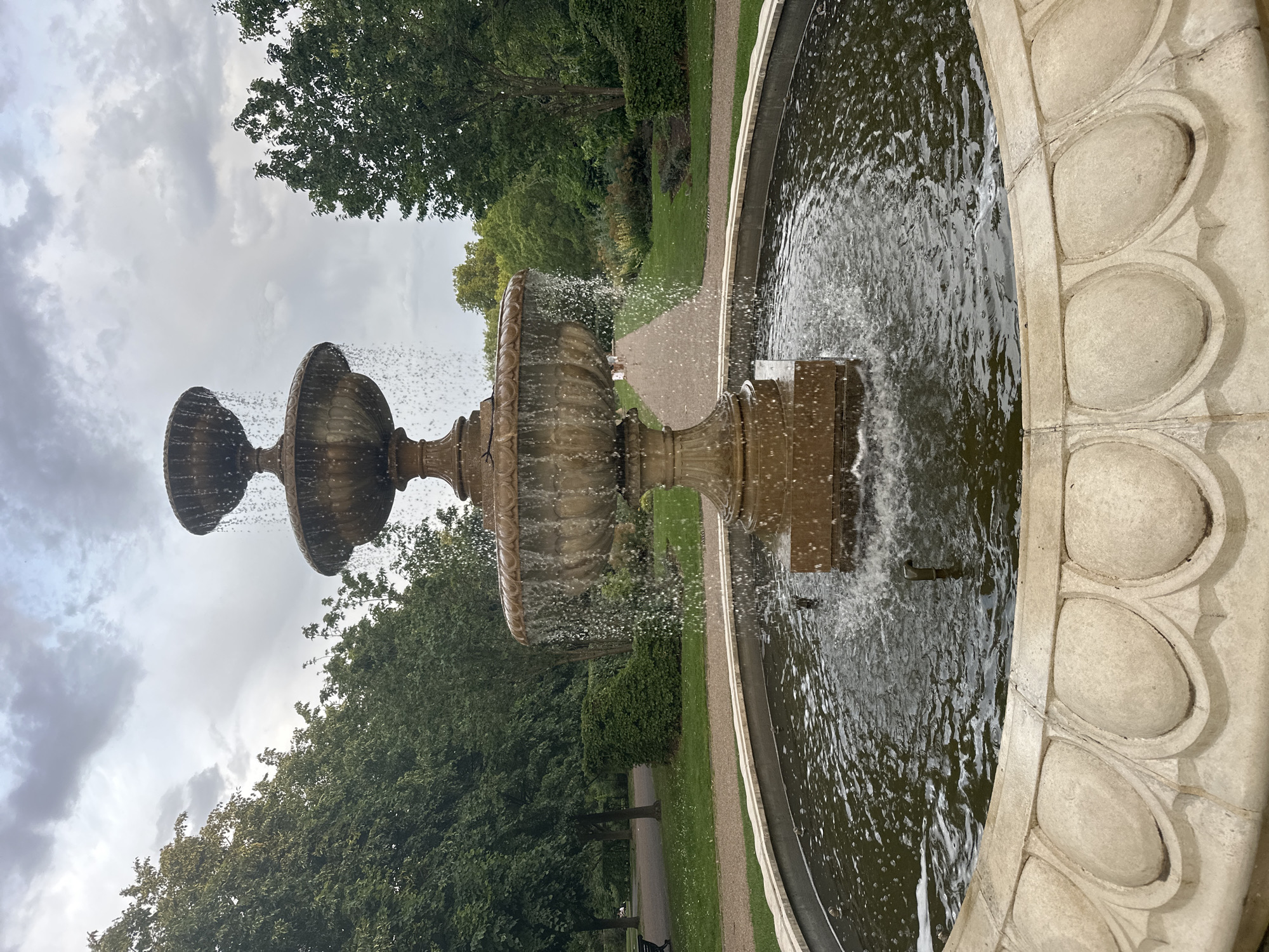 This outdoor scene shows a tiered stone fountain in a circular basin. Water is actively flowing from the fountain into the basin, creating droplets in the air. The basin has a decorative rim with oval-shaped indentations. The background features greenery, including trees and grass, suggesting a park or garden setting. A paved path is visible behind the fountain. The sky is overcast with grey clouds. It depicts a typical view of the Italian Gardens in Hyde Park, London, United Kingdom.