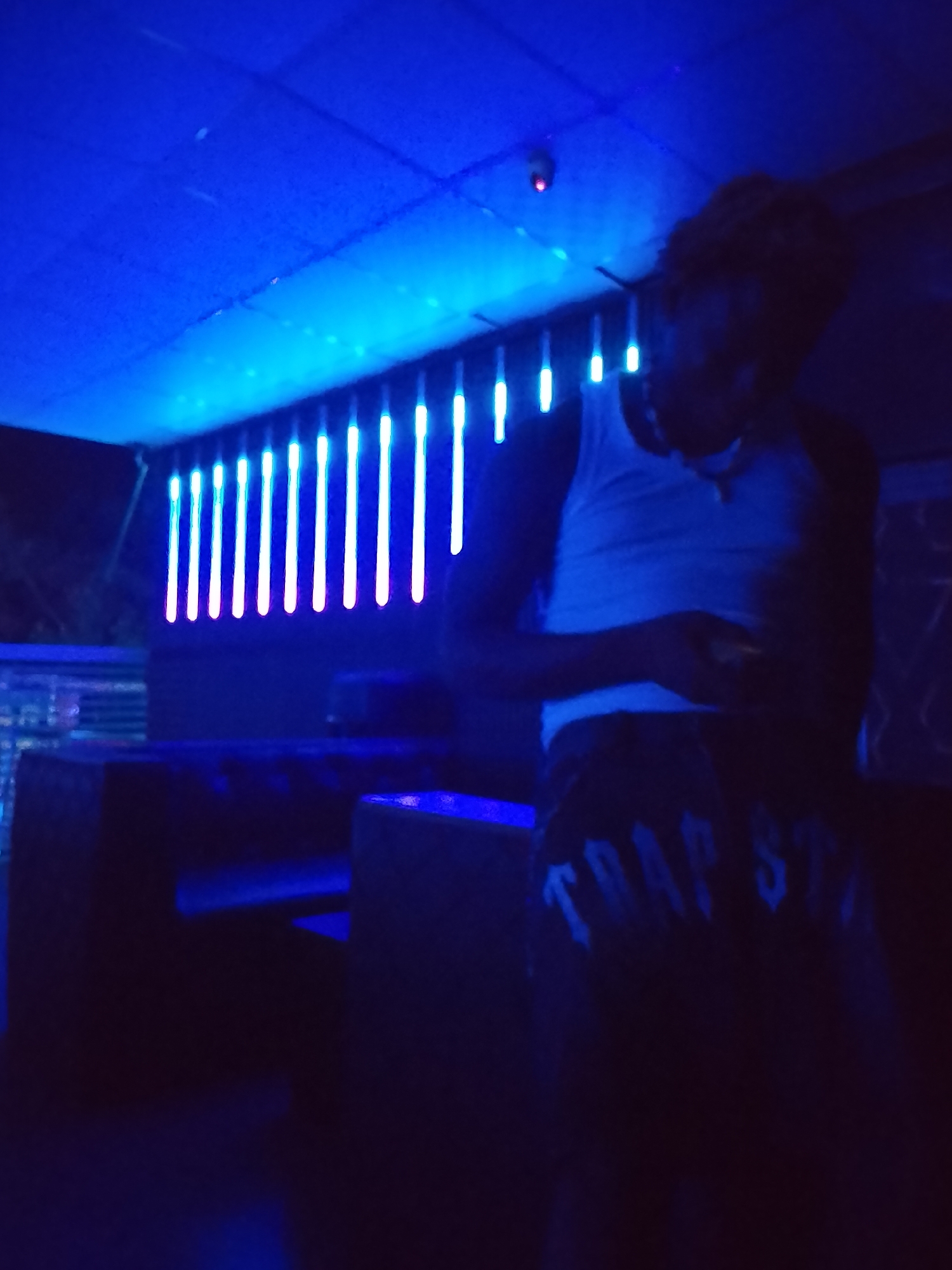 A person wearing a white tank top and dark pants with "TRAP STAR" printed on them stands indoors. The location is dimly lit with a bluish hue. A row of bright vertical lights is visible in the background, likely illuminated by neon lights. Furniture is also present, suggesting the scene is set in a bar. The presence of the person looking at their phone, the lights, and furniture indicate that the subject is at leisure. There is no visual confirmation for the exact location of Narayi, Nigeria.