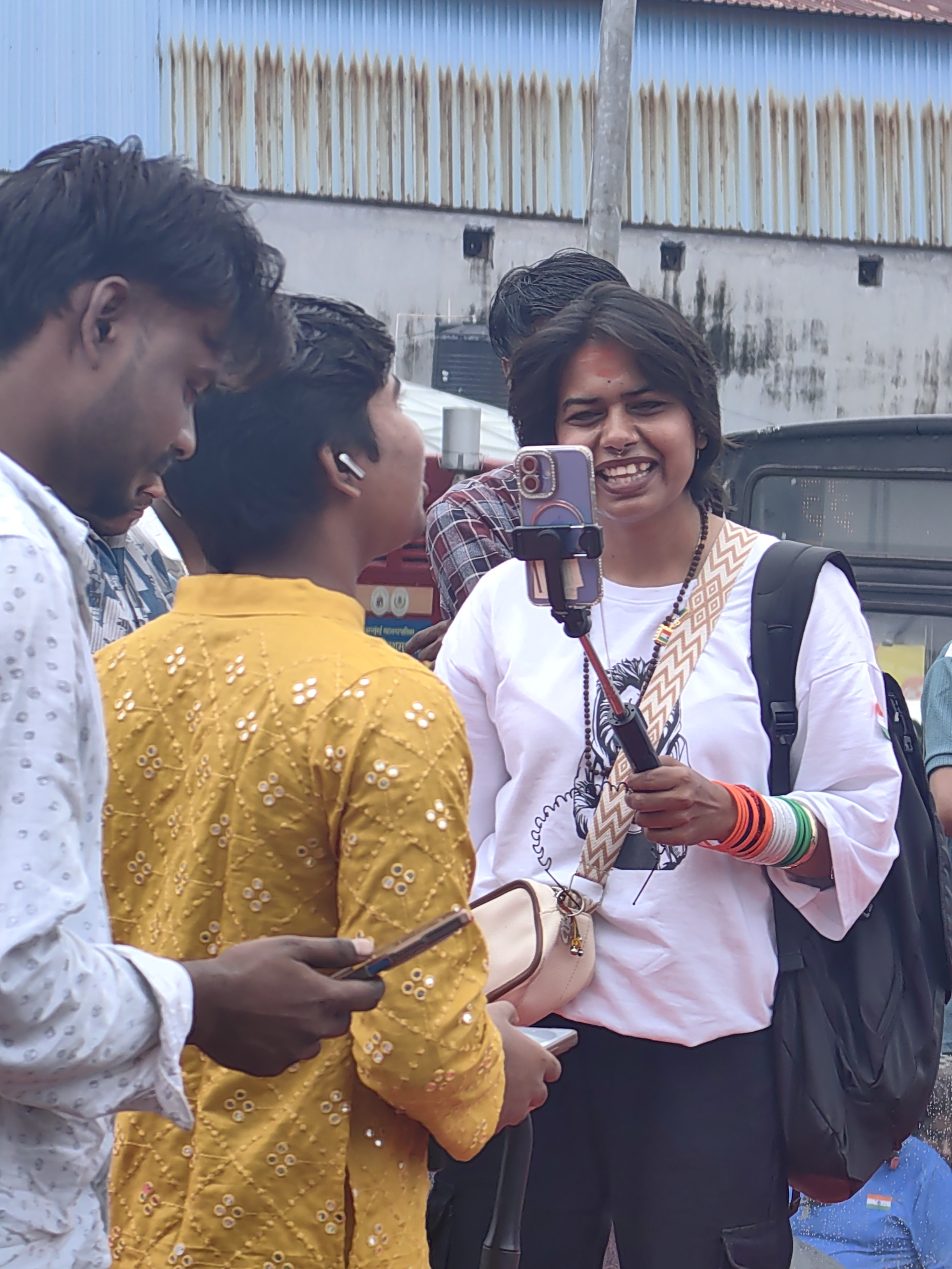 A vertical, medium shot captures a group of young people in a street scene, likely in Mumbai, India, given the presence of what appears to be the Indian flag on a t-shirt and bracelets. In the foreground, a young woman with dark hair, wearing a white t-shirt with a graphic print and black cargo pants, smiles broadly as she holds a smartphone mounted on a selfie stick. She is adorned with a traditional red bindi on her forehead and numerous colorful bangles on her wrist, including green, white, and orange, mirroring the Indian flag's colors. A beige crossbody bag rests on her shoulder. To her left, a young man in a white shirt with a subtle pattern looks down at his phone, while another young man, seen from the back, wears a bright yellow kurta embellished with mirrored white circular patterns. Behind them, a vehicle, possibly an SUV, is partially visible. The background consists of a building with a corrugated metal facade and a textured, light-colored wall, suggesting an urban environment. The overall scene is dynamic and informal, indicating a casual gathering or event.