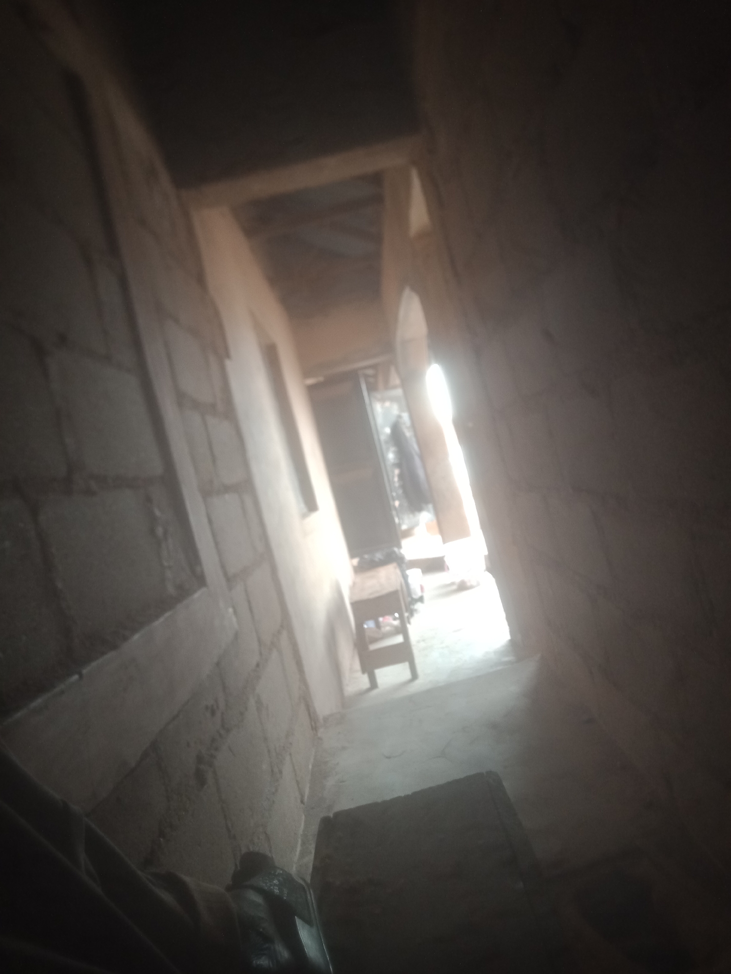 The image shows an interior hallway, possibly under construction or in a state of disrepair. The walls appear to be made of rough, unplastered bricks. There is a narrow window on the left wall, and a bright light source coming from the end of the hallway, suggesting an opening or doorway. In the distance, a small wooden stool or bench is visible. The floor is concrete. The lighting is dim, with strong shadows cast throughout the hallway. The perspective is angled, giving a sense of depth. The foreground is dark and includes what appears to be a person's legs and hand, wearing dark clothing.