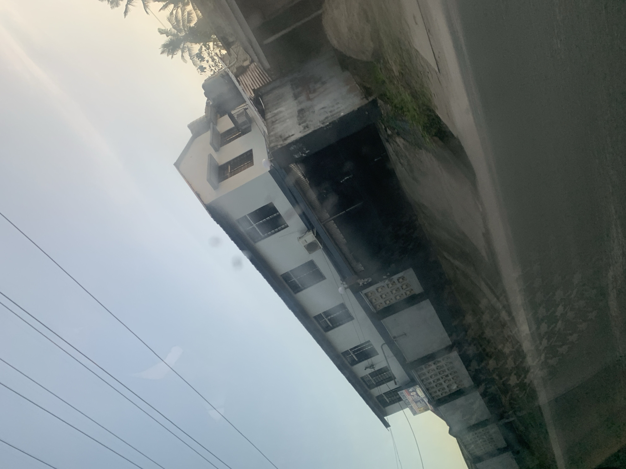 A long building with many windows is visible in the image. A road runs parallel to it. The photograph was likely taken from inside a vehicle. The sky is overcast. There are electrical wires in the foreground. Palm trees are visible in the background. Based on the architecture and surroundings, this image is potentially from Dar es-Salaam, Tanzania.