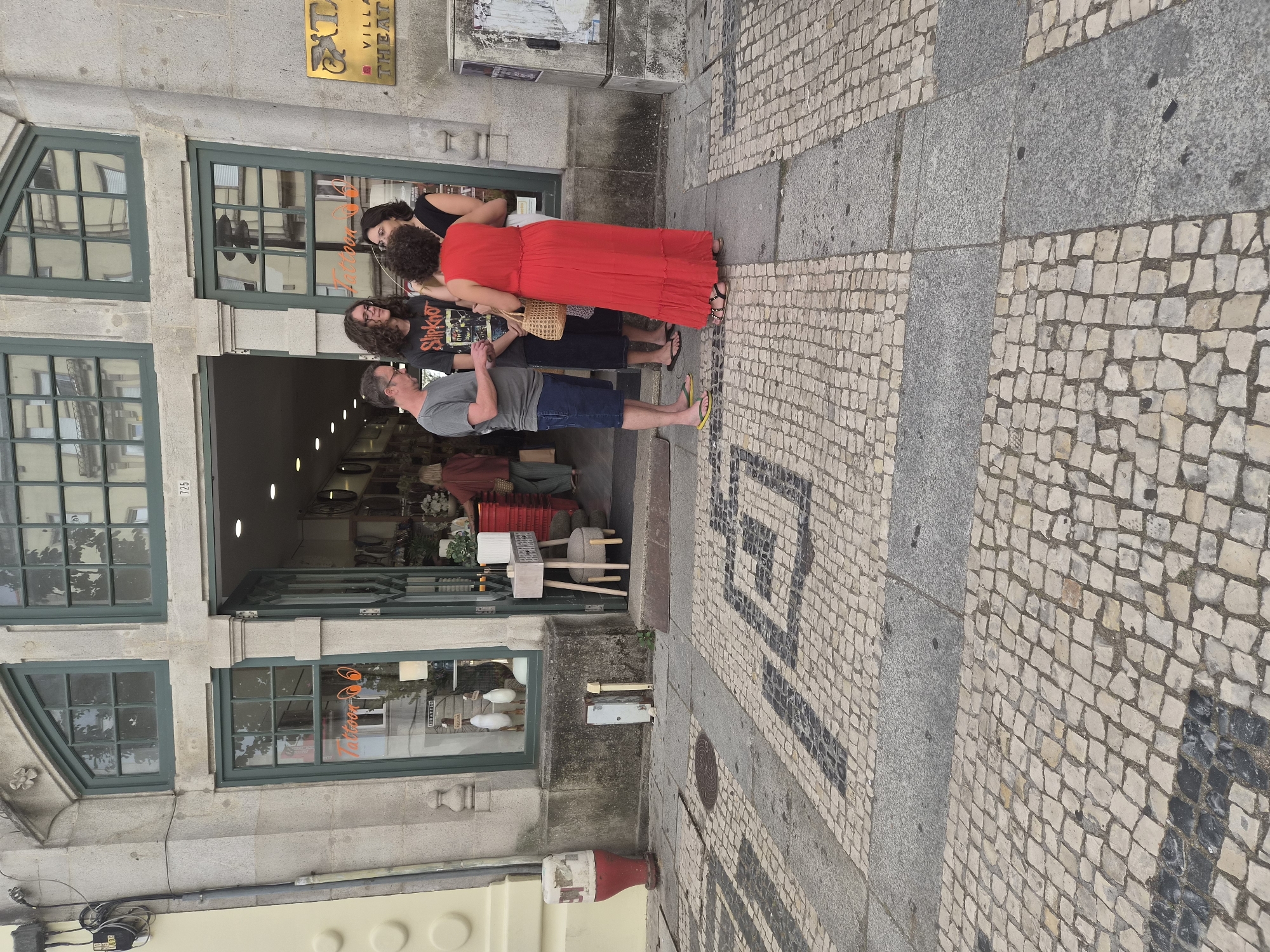 In Braga, Portugal, on a street paved with traditional Portuguese cobblestones, a group of four individuals stands near the entrance of a commercial establishment. The building features a large, arched doorway and windows with dark green frames. Above the entrance, the number "725" is visible. A sign with "Tattoo" in orange letters is displayed on a window to the left. To the right, a golden sign with "EXL" and "VILLA THEATR" is mounted on the building's facade.
The group consists of two men and two women. One man, wearing a grey t-shirt and blue shorts, stands on the left, engaged in conversation. Next to him, a younger man in a black t-shirt with "Slipknot" printed on it is also speaking. A woman in a black dress is on the right, facing the man in the grey t-shirt. The fourth person, a woman wearing a long, bright red dress and carrying a woven bag, is the central figure in the group, facing away from the camera and interacting with the others.
The cobblestone pavement in front of the building is patterned with contrasting dark and light stones, forming geometric designs. In the foreground, a portion of the street shows a different, more uniform cobblestone pattern. To the left, near the entrance, a red and white bollard is visible. The overall scene captures a casual gathering on a city street, suggesting a moment of interaction or discussion between the individuals.