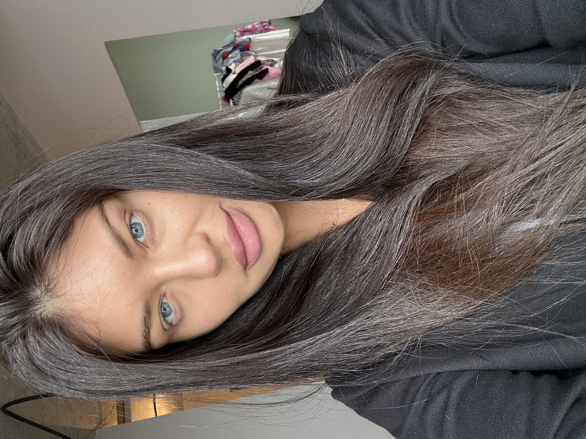 A portrait-style image, rotated 90 degrees clockwise, features a woman with long, dark hair and striking light blue eyes. She is looking directly at the camera with a neutral to subtly smiling expression. Her lips are adorned with a matte pinkish-nude lipstick.
Her hair, which frames her face and cascades over her shoulders, is predominantly dark brown but shows a notable presence of gray and silver strands, particularly visible in the zoomed-in crops near the scalp and throughout the length. A thin silver chain necklace is visible on her neck. She is wearing a dark, possibly black or very dark grey, textured top, which appears to be made of a soft, casual fabric like a hoodie or sweater.
The background suggests an indoor setting, likely a casual home environment. Behind her, a plain light-colored wall is visible, with a section of a muted green color in the upper left. A blurred area in the background, to the right of her head, appears to be a pile of clothes or a laundry hamper, further supporting the relaxed home context. No specific location cues definitively point to Khimki, Russia, other than the provided context. There is no visible text in the image.