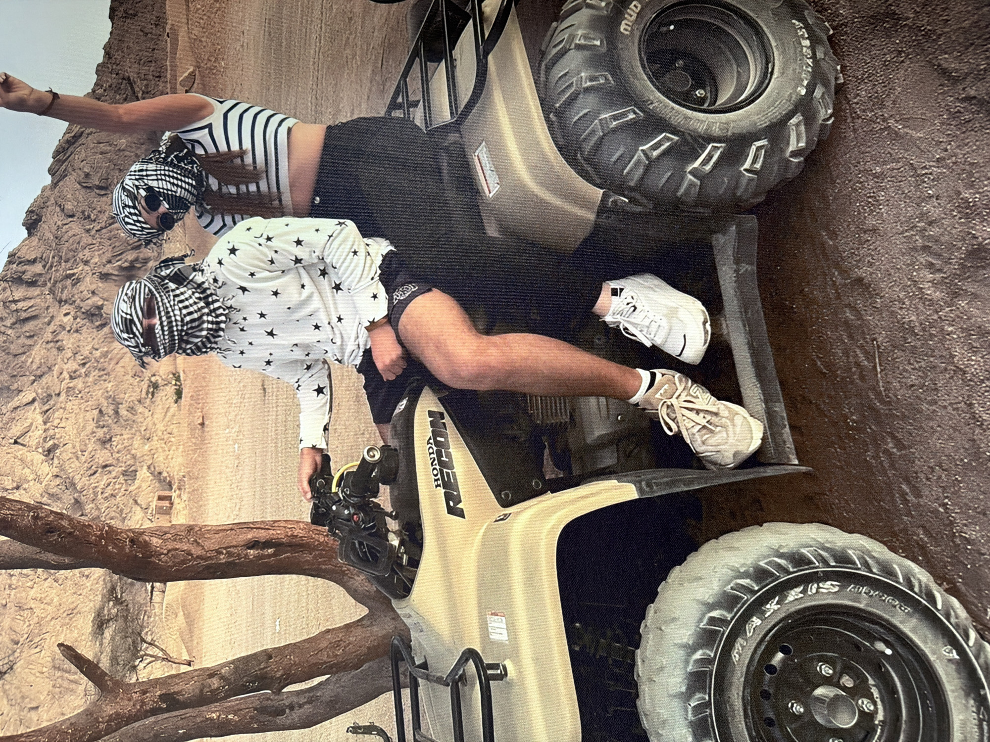 Two individuals are seated on a tan Honda Recon ATV in a desert-like environment. The person in the driver's seat is wearing a white long-sleeved shirt with black stars, black shorts, and white sneakers. They are also wearing a black and white keffiyeh covering their head and face. The second person, seated behind the driver, is wearing a striped crop top and black pants, also with a keffiyeh covering their head and face. Their right arm is raised in the air. The ATV has a black rack on the back. The background features sparse trees with rough bark, sandy terrain, and rocky hills. The image does not provide any information suggesting a location in Strășeni, Moldova.