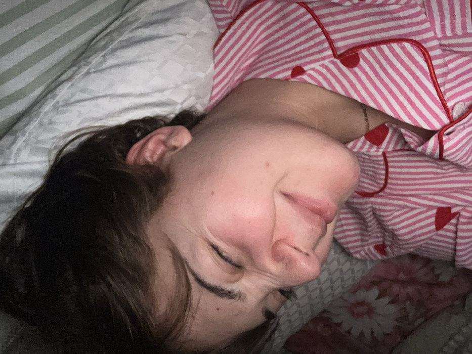A woman is sleeping on a pillow with a striped case. She is wearing pink and white striped pajamas with red accents. Her eyes are closed, and her face is relaxed. Her dark hair is slightly disheveled. Part of a floral patterned blanket is visible beneath her. The scene is dimly lit.