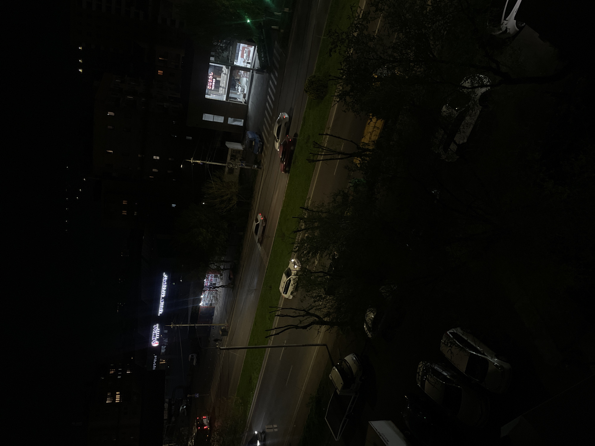 This is an aerial night shot of Yerevan, Armenia. The main street runs diagonally through the frame, lined with buildings. Several vehicles are moving along the street, with some parked on the sides. Trees partially obscure the view. Building lights and streetlights illuminate the scene. Signage in Armenian script is visible on some buildings.