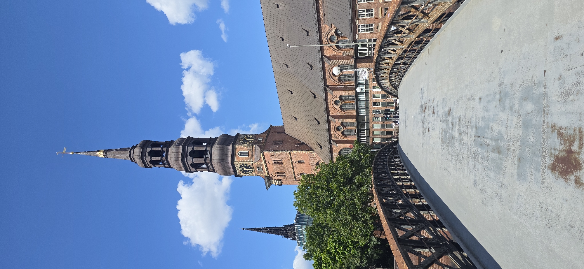 This is a vertical, eye-level, full shot of the St. Nikolai Memorial in Hamburg, Germany. The memorial's brick tower, with a dark, multi-tiered spire topped with a golden cross, dominates the right side of the frame. The tower features arched windows and clocks. A portion of the memorial's copper-colored roof is visible. A dark, ornate bridge with a wrought-iron railing stretches across the foreground, leading towards the memorial. The bridge surface is weathered concrete with rust stains. Green trees are visible to the left of the memorial. In the distance, behind the memorial and trees, another tall, dark spire is visible, likely part of another church or building, partially obscured by a modern glass building. The sky is a clear, bright blue with scattered white cumulus clouds. No people are clearly visible in the image. The scene appears to be a sunny day.
