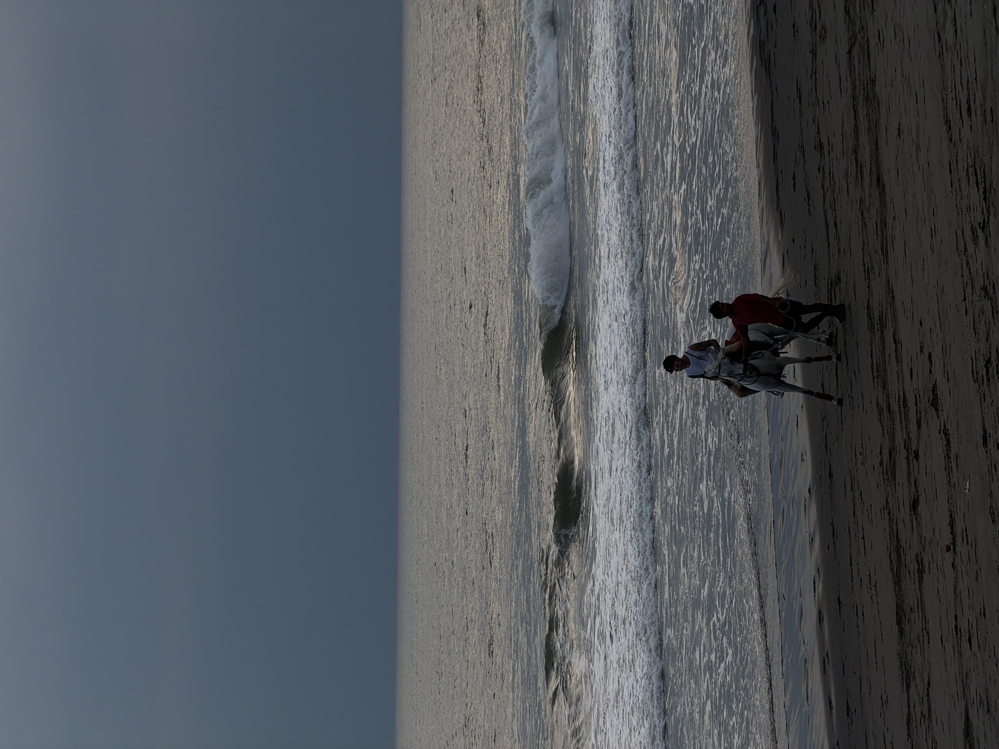 A forensic examination of the image reveals a beach scene in Belalang, Indonesia, under an overcast, dark gray sky. The dark, wet sand occupies the foreground and midground, leading to a dark ocean with multiple white-capped waves breaking near the shore. Two adult males and a white horse are present. One male, wearing a light-colored sleeveless top and dark sunglasses, is mounted on the horse. The second male, dressed in a red short-sleeved shirt and dark bottoms, walks directly beside the horse, holding its bridle, indicating he is leading the animal. Both individuals and the horse are positioned near the water's edge and are moving generally parallel to the shoreline. The overall lighting is dim, consistent with dusk or heavily overcast conditions.