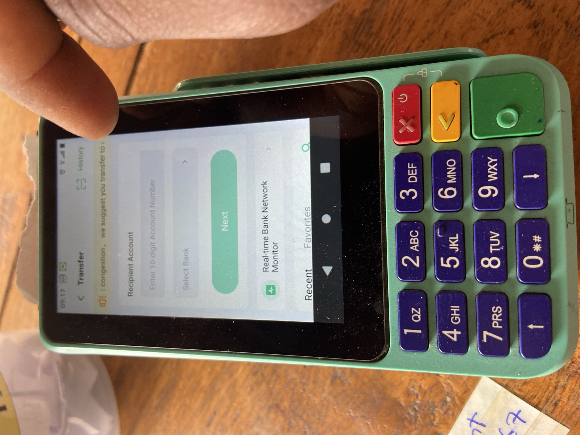 A close-up, top-down, angled outdoor shot shows a green and black point-of-sale (POS) device on a wooden surface. The device has a touchscreen displaying a transfer interface with fields for "Recipient Account" and "Select Bank," and a "Next" button. A finger is obscuring the upper right corner of the screen. The device is surrounded by a wooden surface that appears to be a table. To the left of the POS device, a torn piece of paper with handwritten text is visible. The overall scene is likely in Mararaba, Nigeria, given the context.