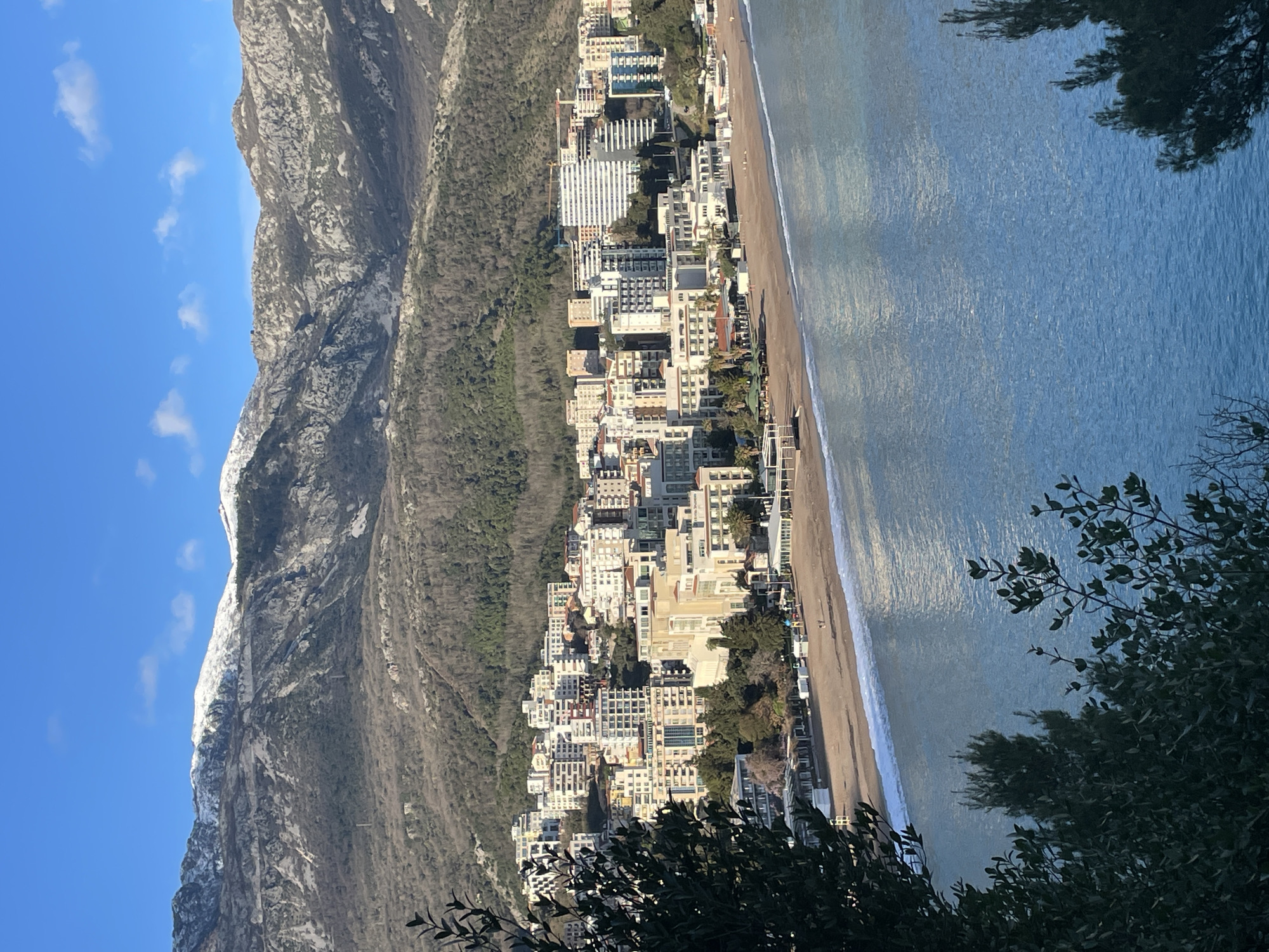 An elevated daytime view captures the city of Budva, Montenegro, focused on Slovenska Plaža (Slovenska Beach) and the urban area expanding inland. In the background, prominent mountains rise steeply, their peaks displaying snow cover, while the lower slopes are characterized by rugged rock formations and sparse deciduous vegetation. The city itself occupies the coastal plain and lower mountain slopes, comprising numerous multi-story buildings, predominantly white, cream, and light gray, consistent with residential and hotel architecture. A construction crane is visible on the right side of the city towards the mountains. Slovenska Plaža, a wide sandy beach, stretches along the coastline in front of the buildings, featuring various small, white and dark-roofed structures, possibly beach bars or changing facilities. Several individuals are discernible walking on the sand. The foreground is dominated by the calm, blue-green waters of the Adriatic Sea, with small waves breaking gently at the shoreline. Dark green foliage from unseen foreground plants partially frames the lower left and right portions of the image. The sky is clear blue with a few scattered white clouds.