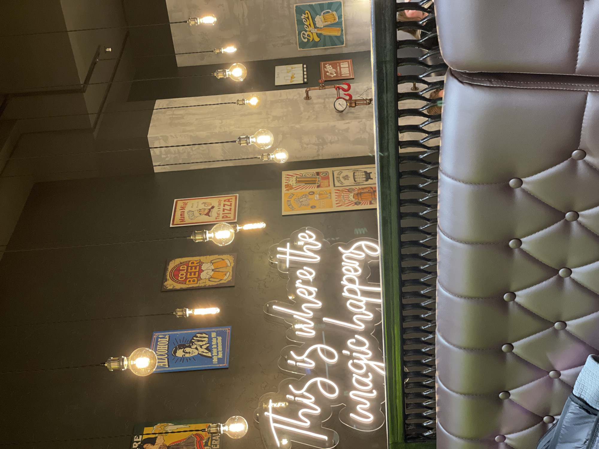 This is an interior shot of a restaurant or bar, taken in a sideways orientation. A tufted, brown, leather booth is on the right. Next to it is a green metal frame. The wall has exposed light bulbs hanging down from the ceiling on cords. There are various vintage-style signs on the wall, including advertisements for pizza, beer, and coffee. There is a neon sign that says "This is where the magic happens". The signs appear to be in English. Given that the OCR of the image recognized "Russia" and "Izhevsk", it is likely that this restaurant is in Izhevsk, Russia.