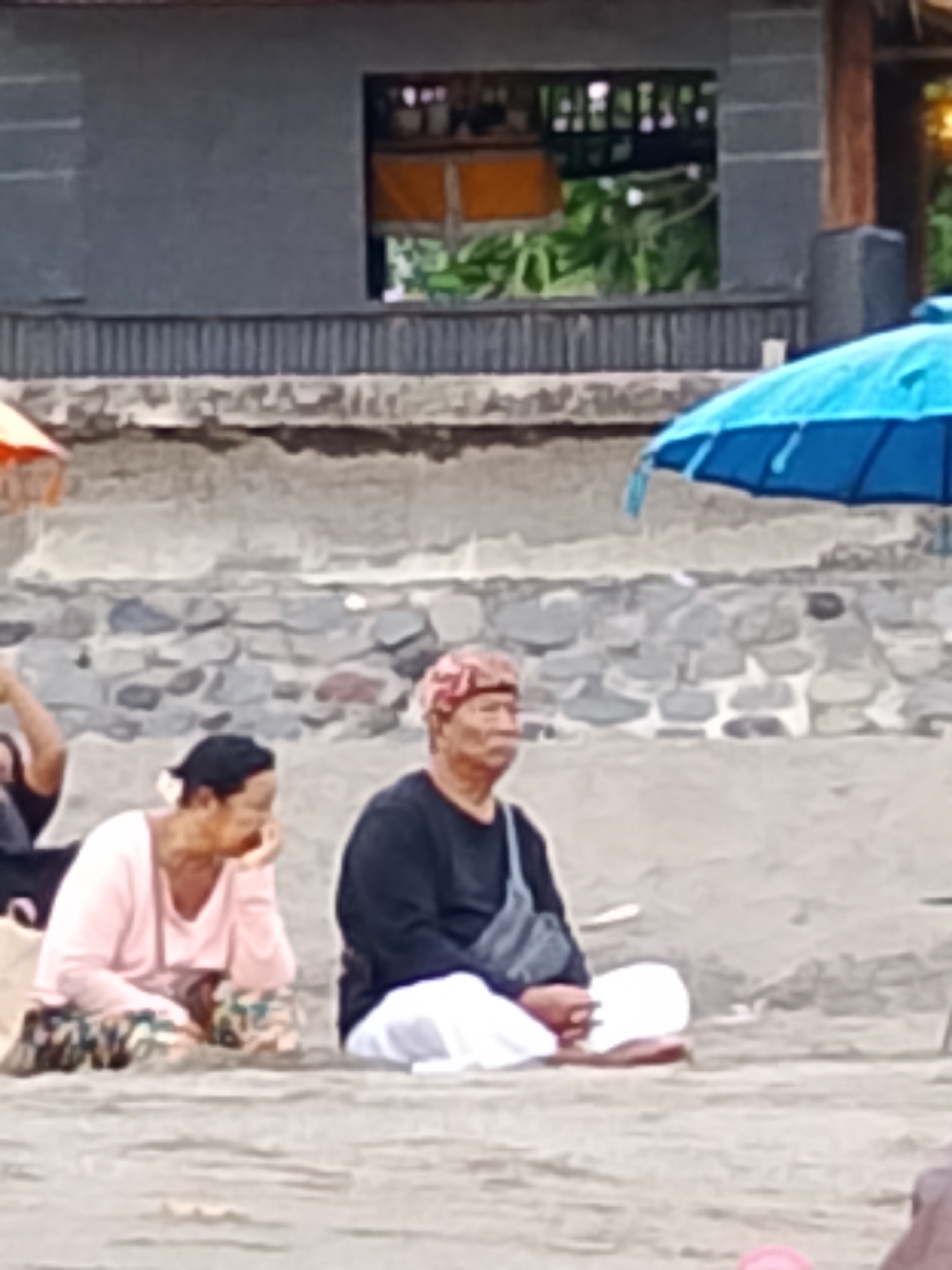The image shows a group of people sitting on a sandy beach in Kediri, Indonesia. In the foreground, a man and a woman are seated. The man wears a dark long-sleeved shirt, white pants, and a patterned headscarf. He has a bag slung across his chest. The woman, to his left, wears a light pink long-sleeved top and patterned pants. She appears to be holding something to her mouth. Behind them, a blue umbrella is visible, partially obscuring a stone wall and a building with a dark gray facade. The building has an open section with shelves displaying various items. The overall scene suggests a casual daytime setting, possibly during a visit to the beach. The lighting indicates it is likely daytime, though the exact time is not discernible. The stone wall and the style of the building could suggest a coastal or resort area. No text is visible in the image.