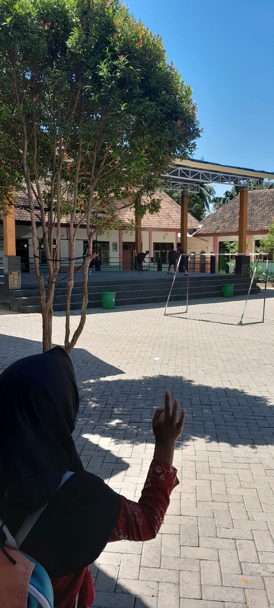 The image depicts an outdoor scene on a sunny day, likely in the city of Lemahbang Dewo, Indonesia. In the foreground, a person wearing a black hijab and a red patterned sleeve is visible from behind, with their right hand raised and fingers extended in a gesture. The ground is paved with light-colored rectangular bricks, and the shadow of a tree with light brown branches and green leaves falls across it.
In the midground, there are steps leading up to a building complex. The buildings have tiled roofs and white walls with pink accents. Several people are visible on the steps and in front of the buildings, some appearing to be interacting with each other. There are also green trash cans placed near the steps.
In the background, a metal structure, possibly a canopy or part of a sports facility, is visible against a clear blue sky. A soccer goal frame is set up on the paved area, suggesting the space might be used for recreational activities. The overall environment appears to be a public area, possibly a school or community center, given the architecture and presence of recreational equipment.