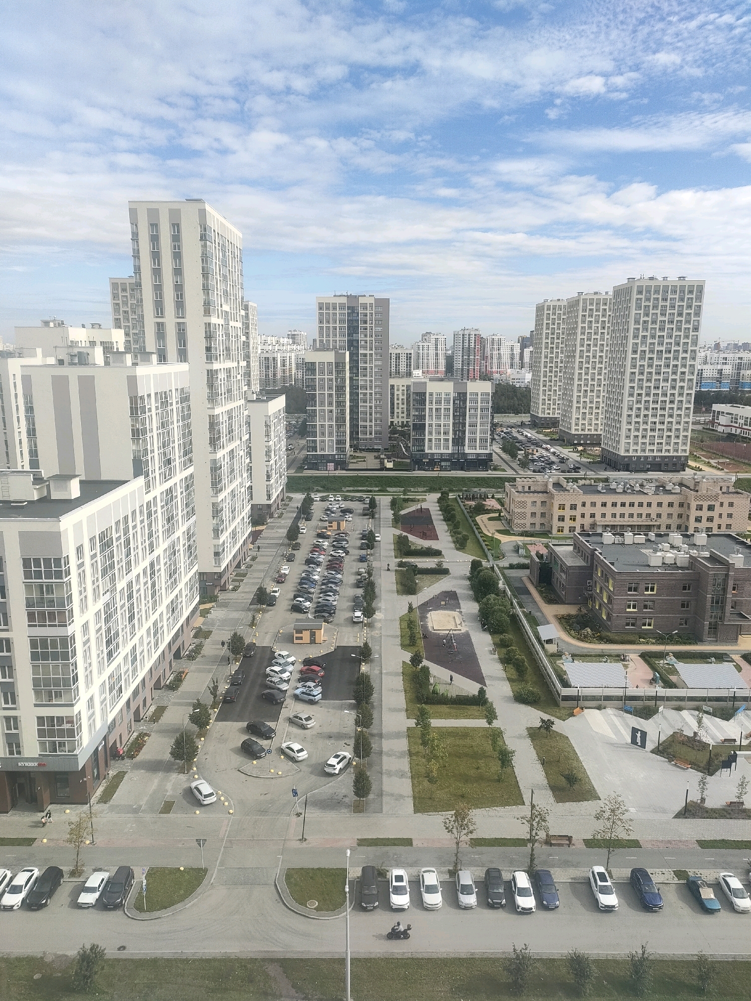 This is a high-angle aerial photograph of a modern residential complex in Yekaterinburg, Russia, on a partly cloudy day. The scene is dominated by numerous multi-story apartment buildings, most of which are light-colored with many windows, characteristic of contemporary urban development. A large parking lot is visible, filled with various cars, and a street runs alongside the buildings, also lined with parked vehicles. In the middle ground, there are a couple of lower-rise buildings, one appearing to be a school or educational institution with an ornate facade. Playgrounds and green spaces with trees and grass are interspersed throughout the complex, indicating a planned community design. The sky above is a pale blue with scattered white clouds. One individual is seen walking on a sidewalk, and another is riding a motorcycle. The overall impression is one of a densely populated, organized urban environment.