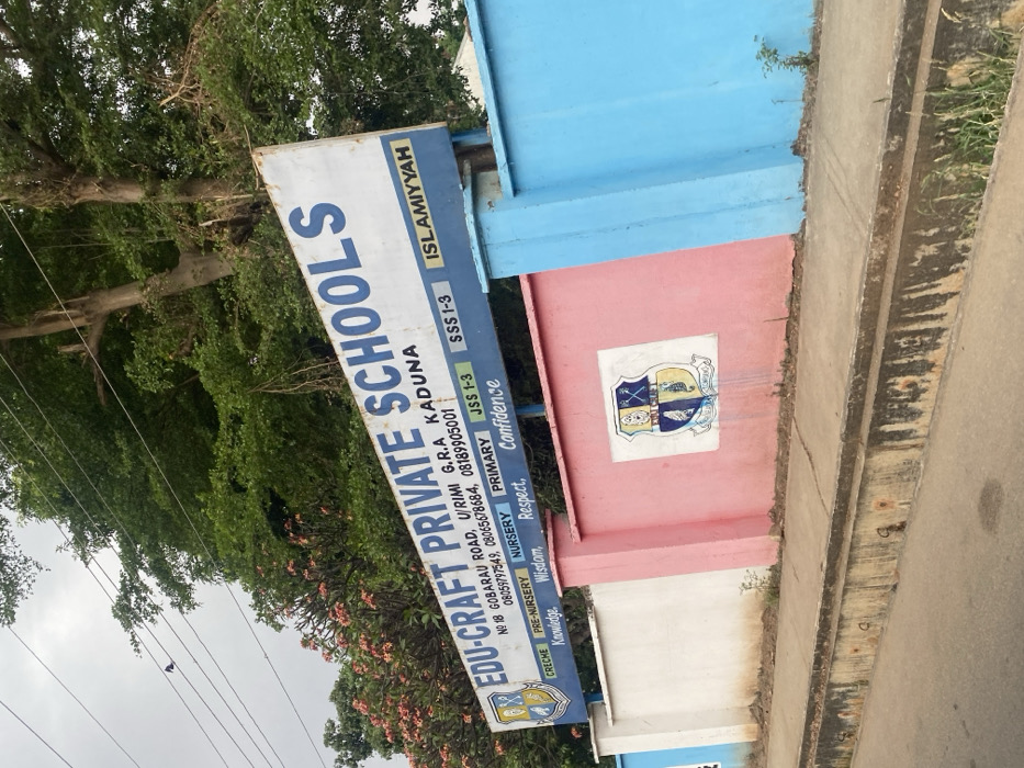 The image depicts the exterior of "EDU-CRAFT PRIVATE SCHOOLS" located at NO 18 GOBARAU ROAD, U/RIMI G.R.A KADUNA, Nigeria. This scene is presented in the context of the city of Rido, Nigeria.
A large, weathered rectangular signboard with a white background and prominent blue lettering displays the school's name, address, contact numbers (08059797549, 0806578684, 08189905001), and lists educational stages: CRECHE, PRE-NURSERY, NURSERY, PRIMARY, JSS 1-3, SSS 1-3, and ISLAMIYYAH. Below these, values such as Knowledge, Wisdom, Respect, and Confidence are noted. A school crest, a shield featuring an open book and crossed implements, is present on the left side of the sign.
Below the signboard, a low perimeter wall runs parallel to the roadside. This wall is sectioned and painted in three distinct colors: white on the left, pink in the center, and light blue on the right. A faded rendition of the school crest is painted on the pink section of the wall. The base of the wall and the narrow concrete sidewalk in front of it show signs of dirt and sparse vegetation. In the immediate foreground, a concrete curb and gutter system runs the length of the wall, appearing discolored and worn.
The upper portion of the image reveals dense green foliage from trees, with some flowering plants exhibiting reddish-pink blossoms behind the left side of the sign. Utility wires are visible traversing the upper left quadrant. The sky is overcast. No individuals are present in the depicted scene.