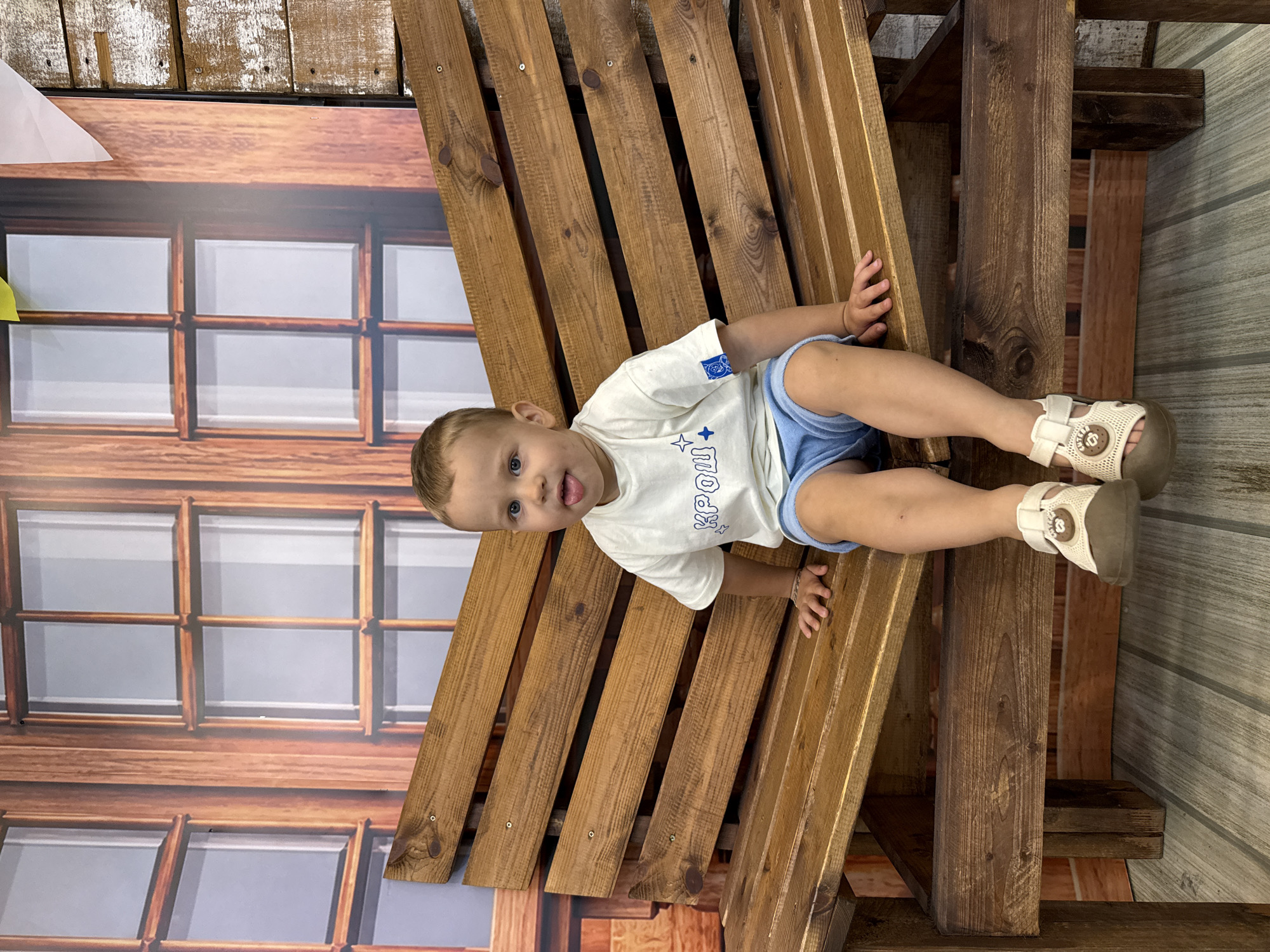 A young boy sits on a wooden bench in front of a backdrop that resembles a wooden house with windows. The boy is wearing a white t-shirt with blue lettering and a blue drawstring shorts. He is also wearing white and brown sandals. His tongue is sticking out. The backdrop features a wooden wall on the right side. The location is not specified as Rusanovo, Russia.