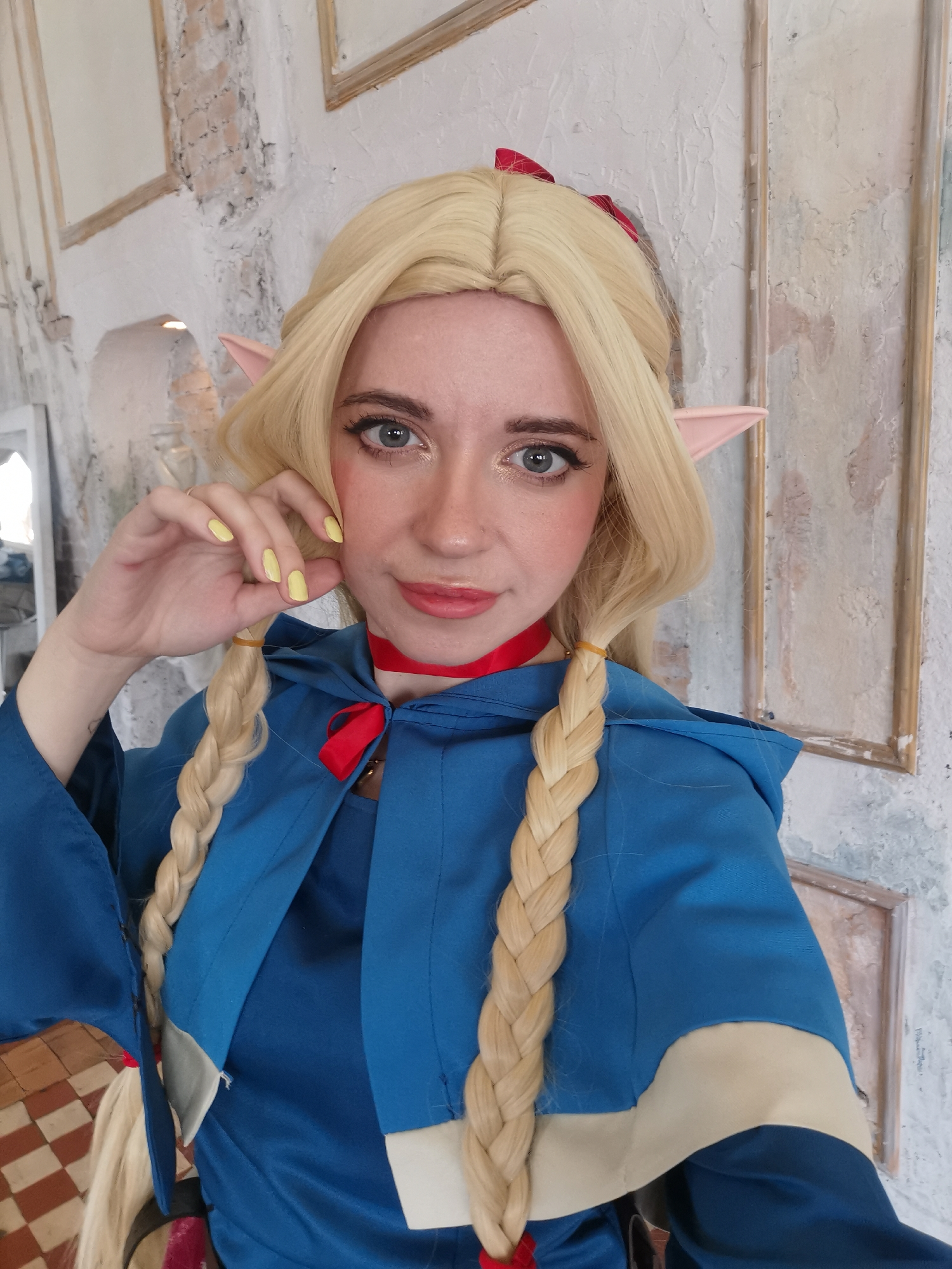 A young woman with fair skin and grey eyes is posing indoors, likely for a selfie or portrait. She is dressed in what appears to be a costume, featuring a blue hooded cloak-like garment with tan trim. Her hair is blonde, styled in two long braids. She also has elf ears and a red ribbon in her hair, and wears a red ribbon as a necklace.
Her left hand is raised to her face, with yellow nail polish visible. The background shows a white wall with framed decorations, and flooring with a checkered pattern.
There is no evidence this is in Moscow.