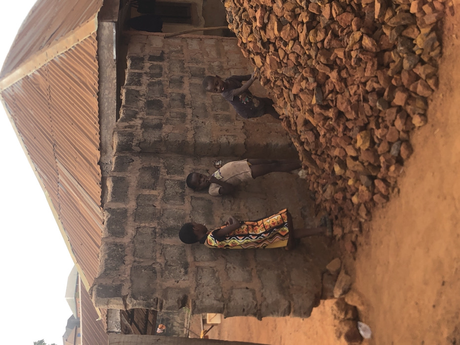 The image portrays an outdoor scene in Nsugbe, Nigeria, featuring three children adjacent to a building and a large pile of rocks. The ground consists of reddish-brown dirt. A building with an unrendered concrete block wall is situated on the right, topped by a weathered, reddish-brown corrugated metal roof. Another structure with a reddish roof is visible in the background, further left.
Three individuals identified as children are present. On the left, a child wearing a multi-colored patterned dress stands on the dirt ground, facing right, with her left hand raised towards her mouth. In the center, a child in a light-colored t-shirt leans against the concrete block wall, holding a small, light-colored object to their mouth. To the right, a third child, dressed in a dark t-shirt, stands near and partially on a large pile of reddish-brown rocks, with one hand resting on the rocks. A piece of white plastic debris is visible on the ground in the lower-left quadrant.