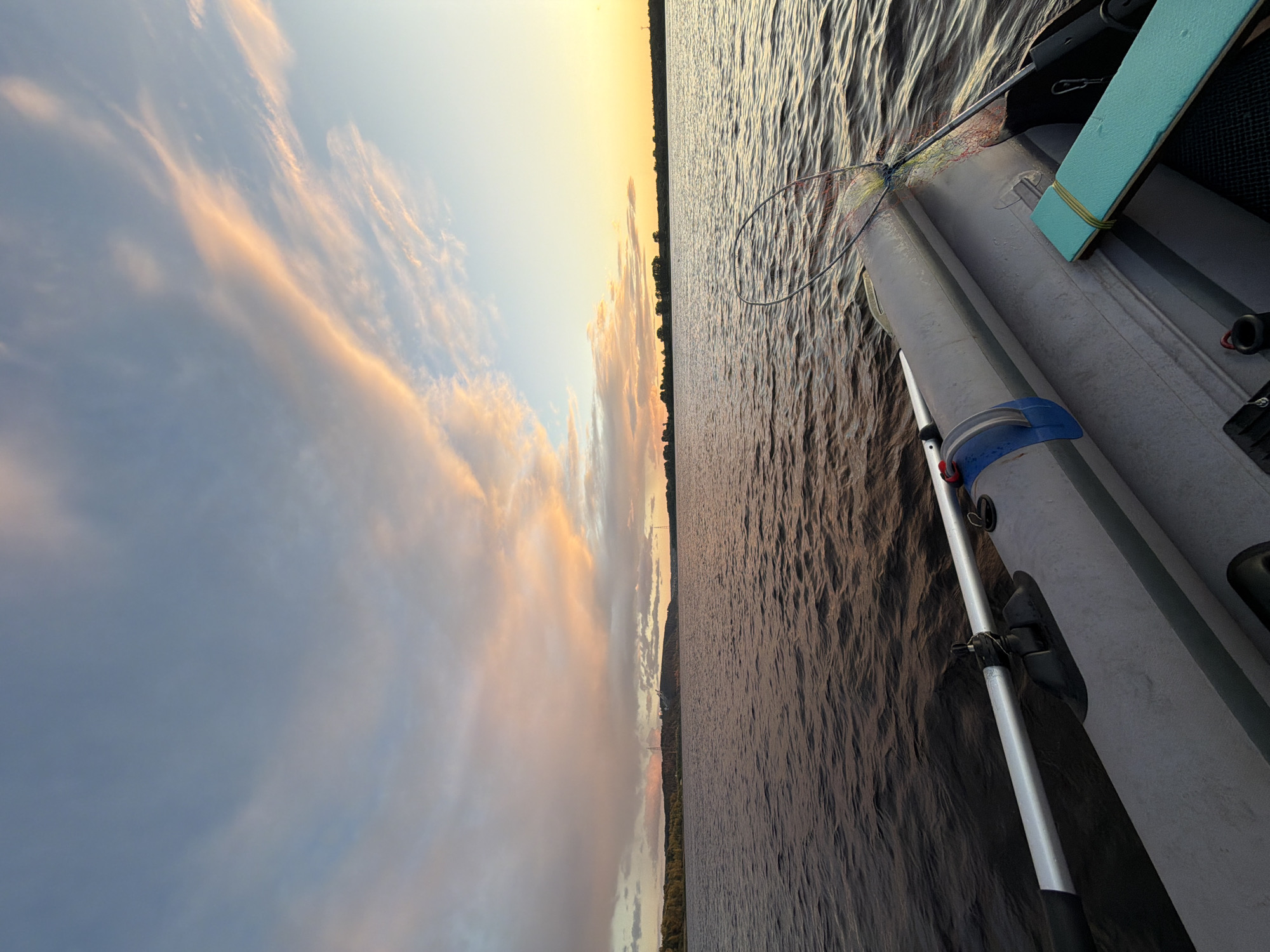 The image depicts a serene water scene at sunset, taken from the perspective of an inflatable boat. The sky is painted with a gradient of soft pinks, oranges, and blues, with wispy clouds stretching across the horizon. The water, appearing calm and dark, reflects the hues of the sky. In the distance, a tree-lined shore is visible, with some structures and what appear to be power lines or transmission towers against the dusky sky.
The foreground of the image is dominated by the interior of the inflatable boat. A silver oar is positioned across the boat, with its handle extending towards the left. The grey, textured material of the boat's inflatable tubes is prominent. On the right side of the boat, a bright turquoise seat or cushion is secured with yellow elastic bands. A fishing net, with colorful threads, is also visible, resting on the edge of the boat. The scene is devoid of any visible people.
The location is the Volga River near the village of Noviy, Russia. The perspective suggests the boat is moving slowly or is at rest, allowing for observation of the sunset and the surrounding landscape. The overall atmosphere is peaceful and tranquil, characteristic of a quiet evening on the water.