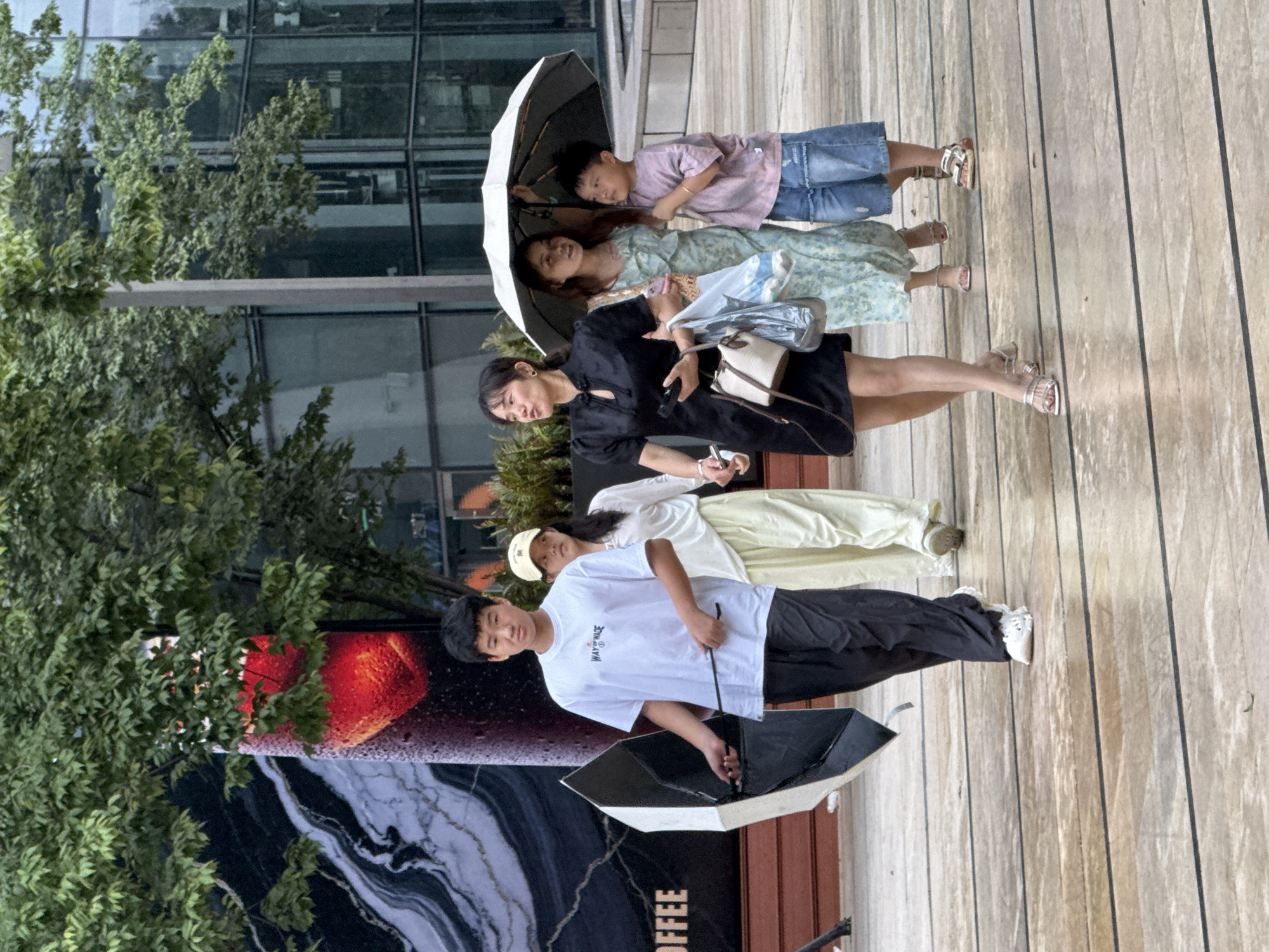 In Yuzhong District, China, on a wet, paved outdoor area resembling a plaza or pedestrian walkway with dark lines demarcating sections of the surface, five individuals are walking. A young male on the left, wearing a white t-shirt with text and dark pants, carries a partially open black and white umbrella. To his right, a person in a light-colored outfit and a beige hat walks. Further right, a woman in a black dress holds a phone and a bag, while a woman in a floral dress holds an open umbrella over herself and a young boy in a light purple t-shirt and denim shorts. The background features lush green trees and a modern building with reflective glass windows. A large advertisement with a red abstract design is visible on the left. The ground appears damp, suggesting recent rain.
