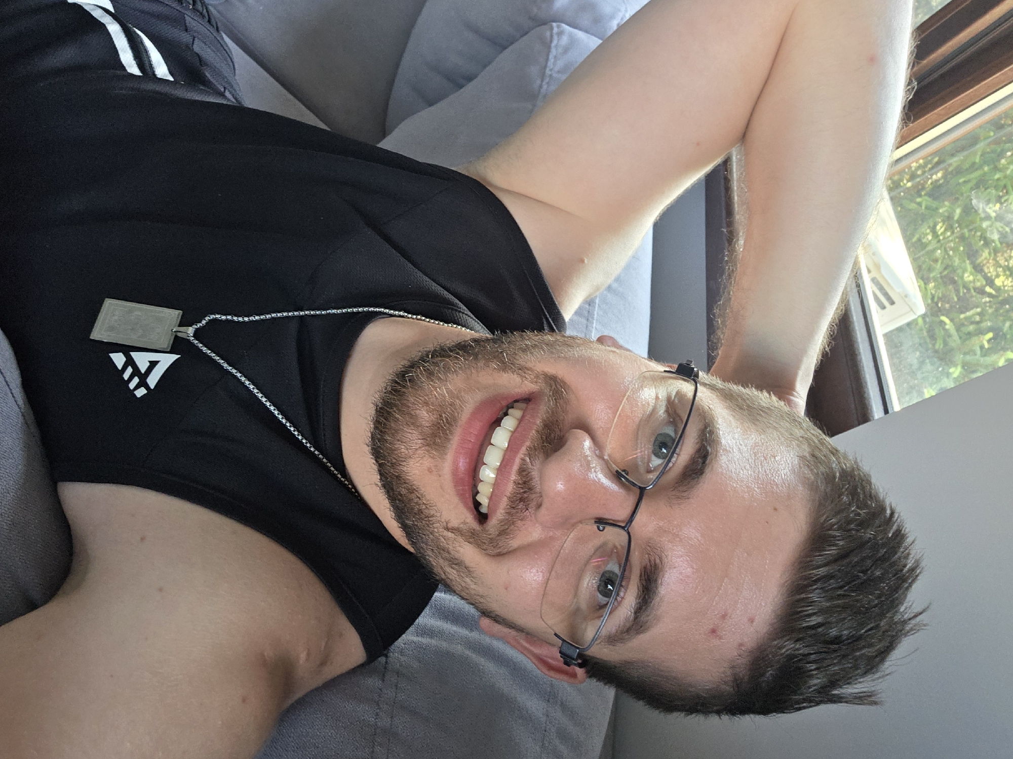A young adult male is captured in a close-up, diagonal selfie, smiling broadly while lying on a light gray sofa or cushion. He is positioned with his head towards the bottom right of the frame, looking upwards directly at the camera. He has short brown hair, a well-trimmed beard and mustache, and fair skin, with visible blue-green eyes behind clear-rimmed glasses. His mouth is open in a wide, relaxed smile, showcasing white teeth.
He is wearing a black sleeveless athletic top, on which a silver chain necklace with a rectangular, metallic grey pendant is visible. The top features a white triangular graphic with a prominent "W" letter on the left side of his chest. In the upper left corner of the image, black athletic shorts with white stripes are partially visible. One of his arms is bent and raised, resting behind his head, while the other is mostly out of frame.
The background shows bright natural light streaming in from a window in the upper right, revealing lush green foliage and trees outside, suggesting a sunny daytime setting. The overall mood is relaxed and happy. The location is indoors, likely a residence in Yagodovo, Bulgaria.