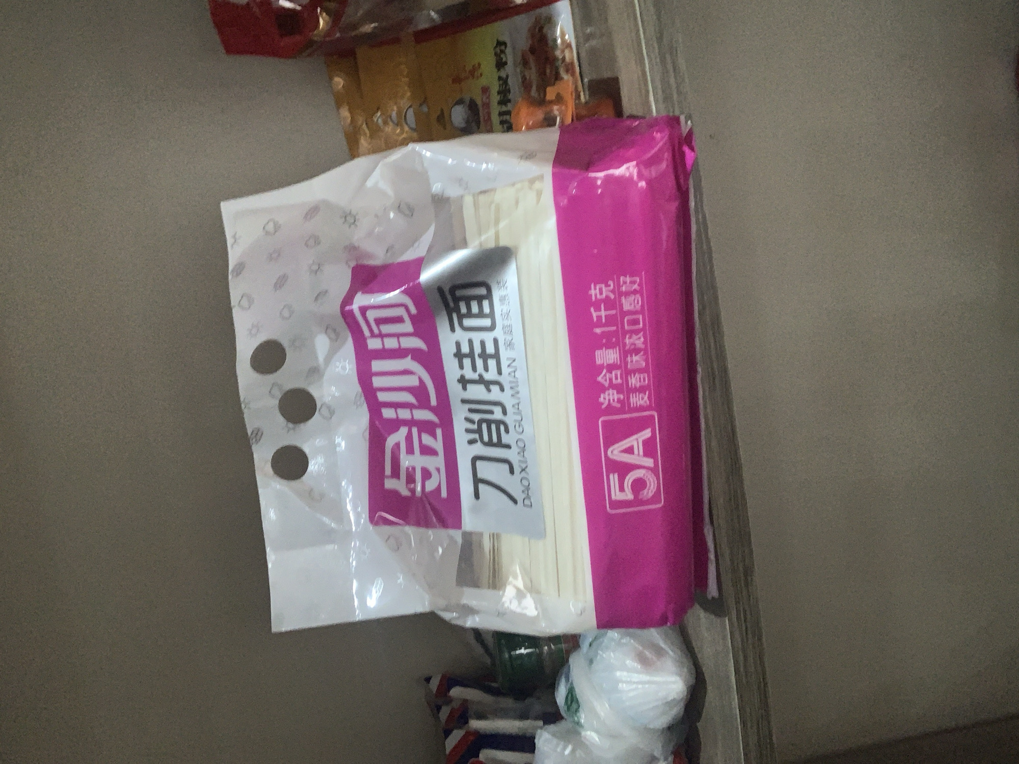 In Abuja, Nigeria, a package of noodles is displayed on a shelf. The package is predominantly pink and white and features Chinese characters and the "5A" logo. The noodles inside are thin and white. To the left of the noodle package, several white plastic-wrapped items are visible. In the background, a yellow package with red and orange lettering is partially obscured. The lighting suggests an indoor environment.