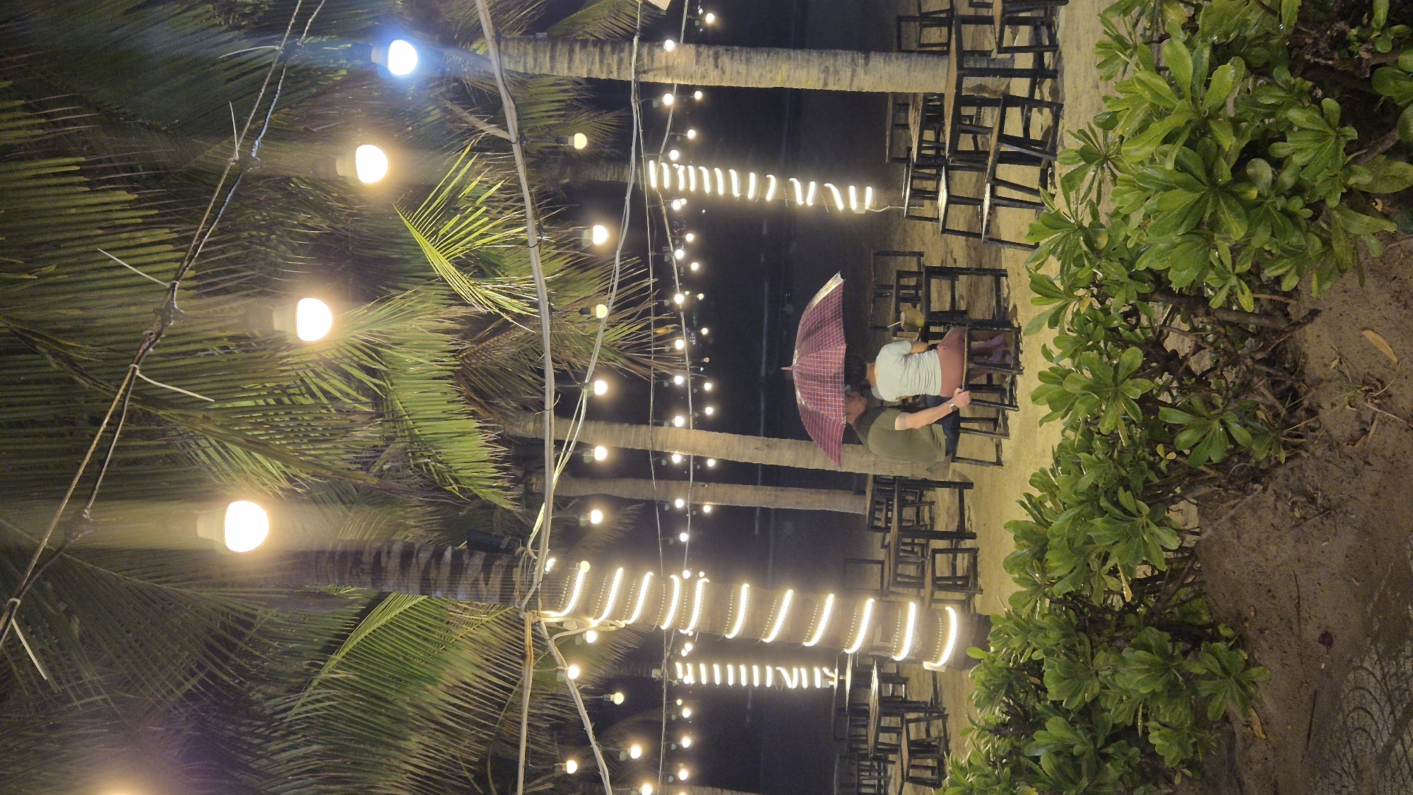 This image depicts an outdoor seating area at night, likely a beachfront restaurant or bar, situated in Đà Nẵng, Vietnam. The scene is illuminated by string lights and lamps suspended among tall palm trees. In the foreground, lush green bushes and sandy ground are visible. In the midground, a couple is seated at a table under a pink umbrella, sheltering from what appears to be rain. They are facing away from the camera. Numerous empty tables and chairs are arranged throughout the area, suggesting a public dining or gathering space. The background reveals the dark expanse of the ocean and the night sky.