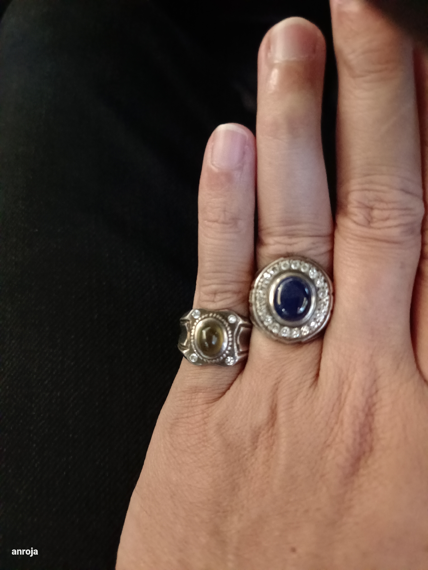 A person's hand, adorned with two rings, rests against a dark, textured background. The ring on the left is silver with a yellowish-green gemstone surrounded by smaller clear stones. The ring on the right is also silver, featuring a large, deep blue gemstone encircled by numerous small clear stones. The hand appears to be resting on dark fabric, possibly clothing, which creates a stark contrast with the skin tone and the shiny jewelry. The lighting highlights the textures of the skin and the facets of the gemstones. The location "Blang Cut, Indonesia" is not discernible from the image itself.