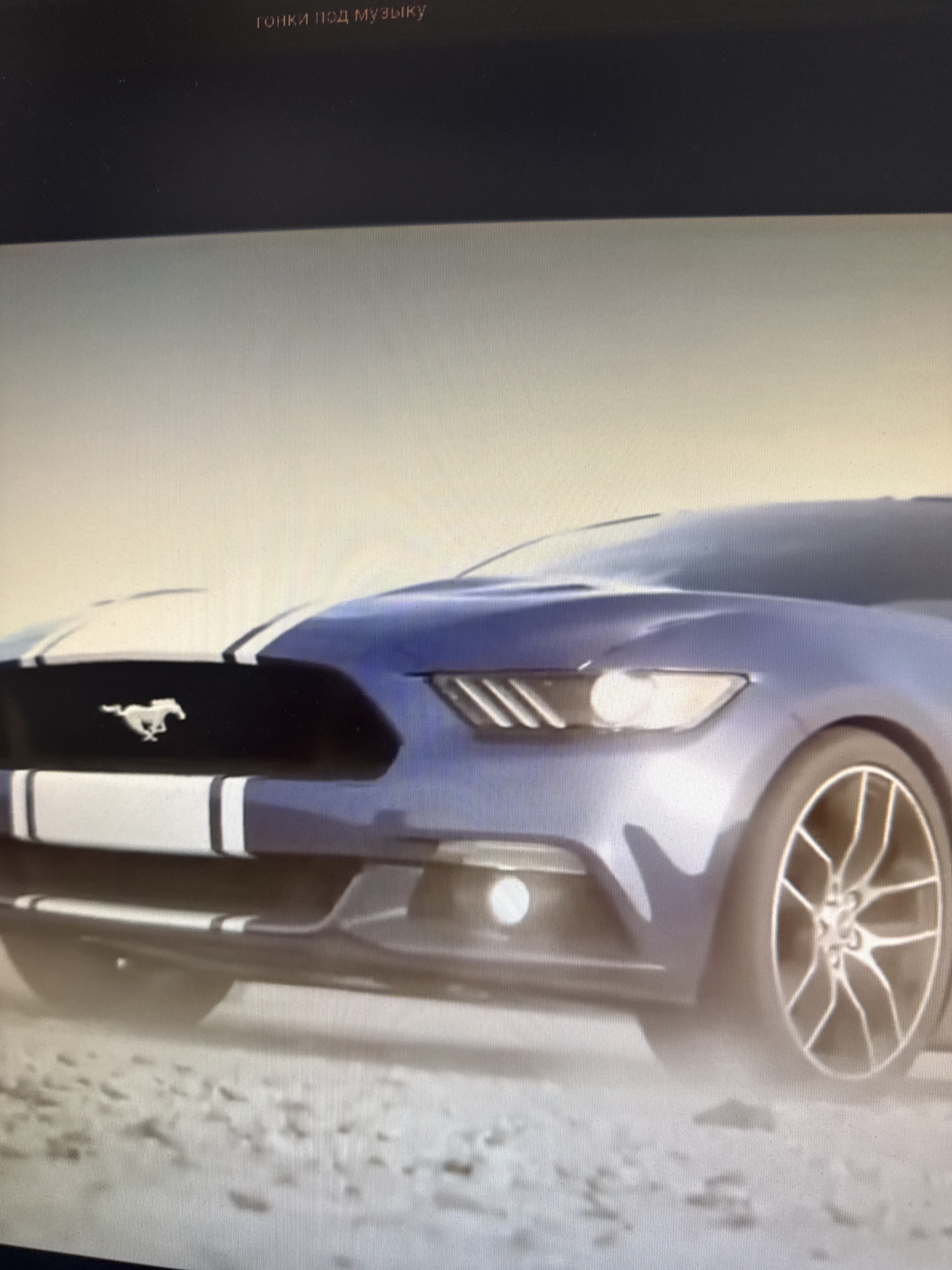 The image shows the front quarter panel of a blue Ford Mustang with white racing stripes. The car is on a sandy or dirt terrain. The background is a light, hazy sky. Text, which appears to be in Russian, is visible at the top of the image, but it is out of focus and too blurry to decipher. The scene is not identifiable as being in Netanya, Israel.