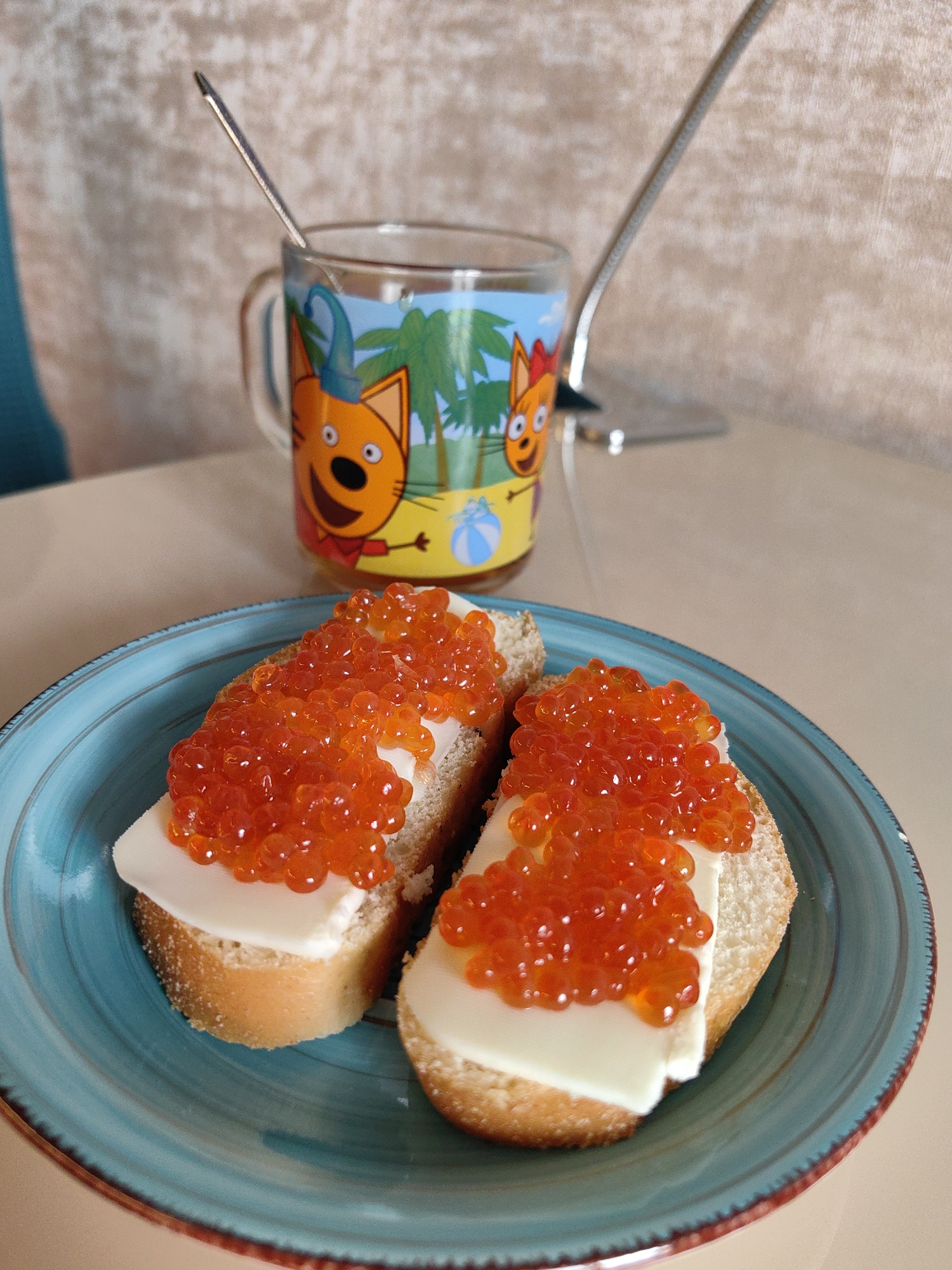 A close-up, eye-level shot shows two slices of bread, each topped with a slice of white cheese and a generous portion of red caviar. The sandwiches rest on a blue, textured ceramic plate with a reddish-brown rim. In the background, a clear glass mug with a cartoon image of two orange cats and palm trees is visible. A silver spoon is placed inside the mug. The background is a light-colored, textured wall and a beige table surface. The scene is lit from the front and slightly to the left. There are no people visible in the image. The setting appears to be an indoor dining area, possibly in Sochi, Russia.