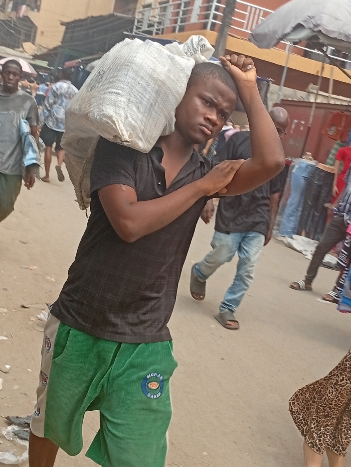 The image portrays an outdoor street or market scene in Fegge, Nigeria, under daylight conditions. A young, dark-skinned male, approximately 18-25 years old, is the central figure, facing slightly left of the viewer. He is dressed in a black short-sleeved polo shirt and green and beige shorts, the latter bearing a circular "MOPAR GARAGE" logo on the left thigh. He carries a large, white, woven sack over his right shoulder. His left hand is raised, resting on the back of his head/neck, while his right forearm is bent across his body. The ground is a light-colored, unpaved surface with some debris. In the background, multiple pedestrians are visible, along with market stalls or buildings constructed from materials like corrugated metal. An open grey market umbrella is partially visible on the right side. Individuals in the background include a person in a light shirt on the left and another in blue jeans and sandals on the right.