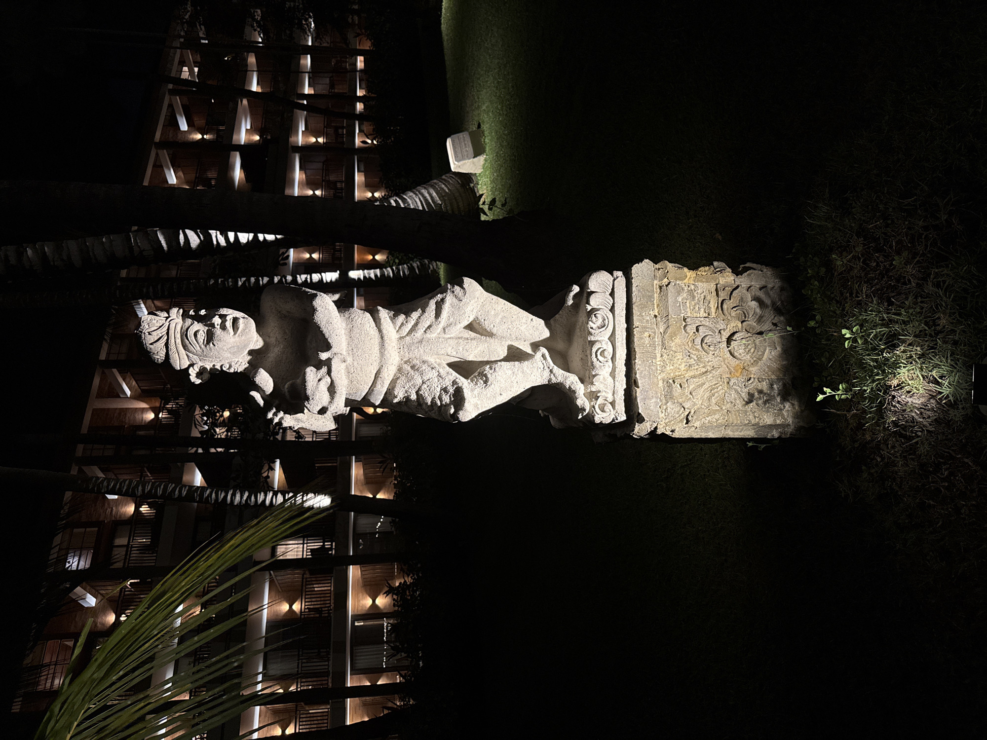 The media file presents an outdoor night scene featuring a large, intricately carved stone statue set within what appears to be a garden or resort. The statue, depicting a traditional figure or deity, stands upright on a decorative base. It has an elaborate headdress, detailed facial features, and is adorned with intricate traditional attire. Spotlights brightly illuminate the figure and its base, casting deep shadows that emphasize the texture and depth of the carvings. The figure's arms are bent, with hands positioned near the waist/chest area, though specific objects held are indistinct due to the lighting.
In the background, a multi-story building, likely a hotel or resort, is visible to the left of the statue. Its numerous windows and balconies are lit, creating a warm glow against the dark night and revealing a modern architectural style with exposed structural elements. Several dark palm tree trunks are visible, framing the scene vertically. To the right of the statue and in the foreground, patches of dark ground are interspersed with illuminated grass and bushy foliage. A large palm frond partially obscures the lower-left section of the building. The overall ambiance is dark, characteristic of nighttime, with artificial lighting strategically highlighting the main subjects and creating strong contrasts. A small, light-colored rectangular plaque or sign is visible in the grass behind the statue, but no text on it is discernible. The style of the statue and the presence of palm trees, combined with the location context of Kuta Selatan, Indonesia, suggest a Balinese or Indonesian cultural setting.