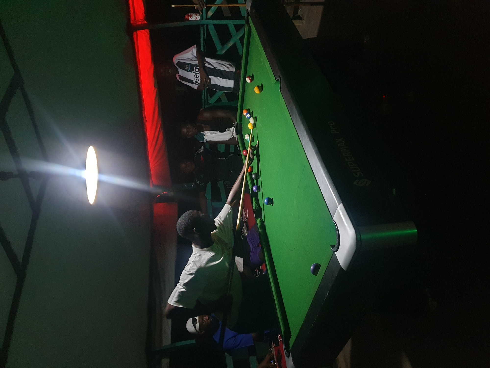 The image depicts a group of individuals gathered around a pool table in an outdoor or semi-outdoor setting, likely in Lokoja, Nigeria, given the context of the request. The scene is dimly lit by an overhead lamp, casting a warm glow on the green felt of the pool table and the surrounding area. Several people are visible, some actively engaged in playing pool while others observe. One person is in the process of taking a shot, cue stick in hand, focused on the balls on the table. Another person, wearing a striped jersey, stands in the background, observing the game. The environment appears to be a recreational space, possibly a bar or a private gathering area, with a canopy overhead supported by green painted wooden structures. The pool table itself is a standard size, with various colored balls arranged on its surface. The overall impression is one of a casual social gathering centered around a game of pool.