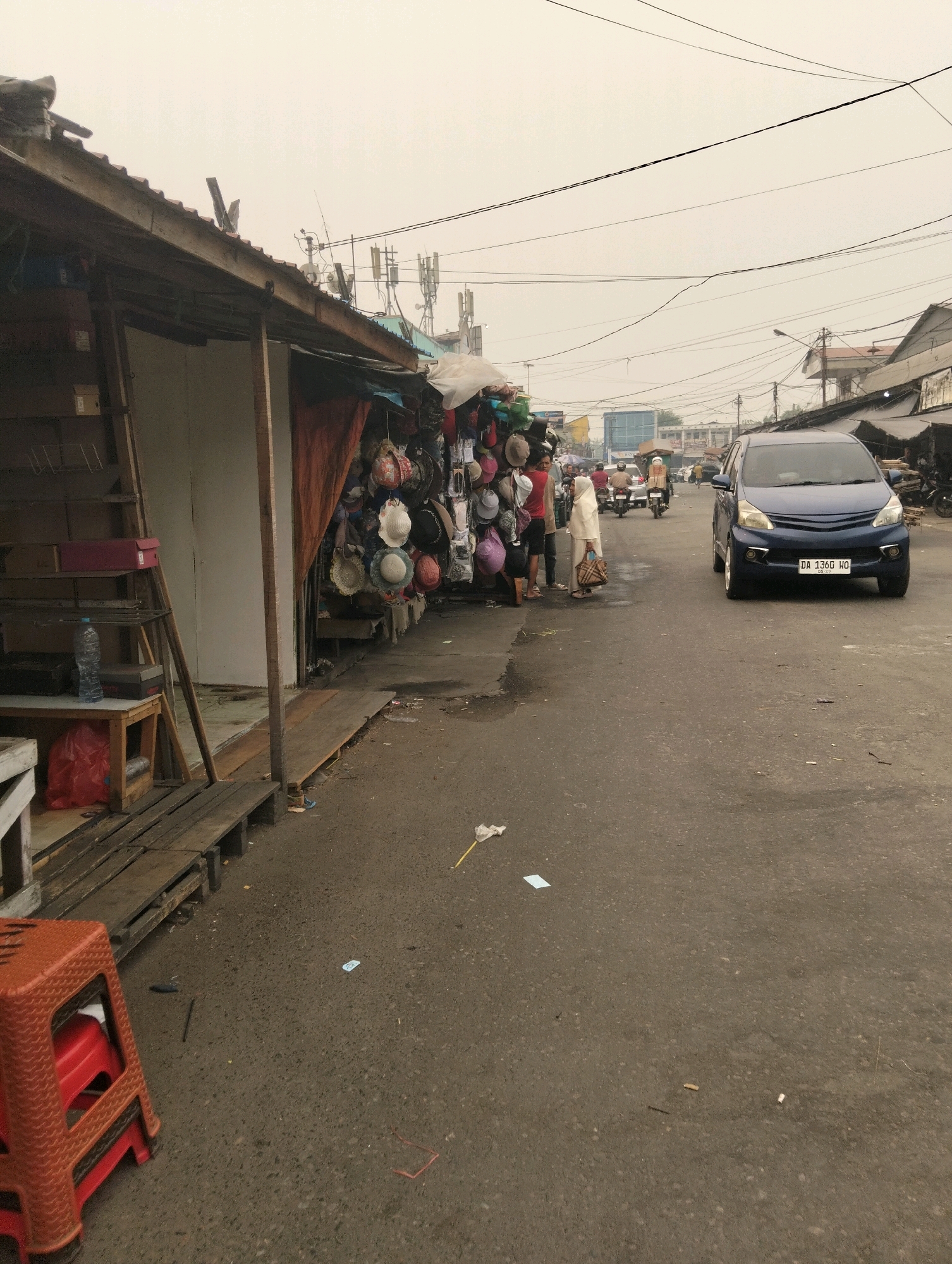 The image depicts a street scene in Palangka Raya, Indonesia, likely a market area. A dark blue minivan with license plate "DA 1360 HO" is parked on the right side of the asphalt road, facing towards the viewer. On the left side of the road, a stall is set up with a wooden awning, displaying various hats for sale. Two individuals, a woman in a white headscarf and a patterned tote bag, and a man in a red t-shirt, are standing near the stall. The street is somewhat littered with debris. In the background, several motorcycles are visible, and more stalls and buildings line the street. Utility poles and wires crisscross the sky, which is overcast. The overall impression is a bustling, outdoor marketplace.