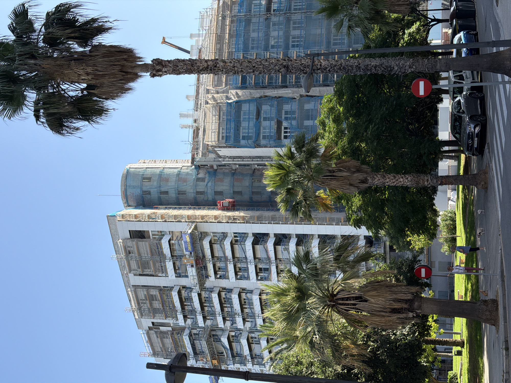 The image depicts a street scene in the city of Valencia, Spain, focusing on a large apartment building undergoing renovation. The building is extensively covered in scaffolding and blue netting, indicating ongoing construction work. A red construction elevator is visible on the side of the building. Several palm trees are prominent in the foreground and mid-ground, with their fronds casting shadows. In the background, a crane is visible.
On the sidewalk in the foreground, a silver Mercedes-Benz SUV and a white van are parked. A blue car is parked further down the street. Two women are standing on the sidewalk with three small dogs. One woman is wearing pink pants and a white top, and the other is wearing a dark blue top and dark pants. A circular red and white traffic sign is posted on a pole. The ground is paved with asphalt and marked with pedestrian crossing lines. The sky is clear and blue. The overall impression is a sunny day in an urban environment with ongoing construction.