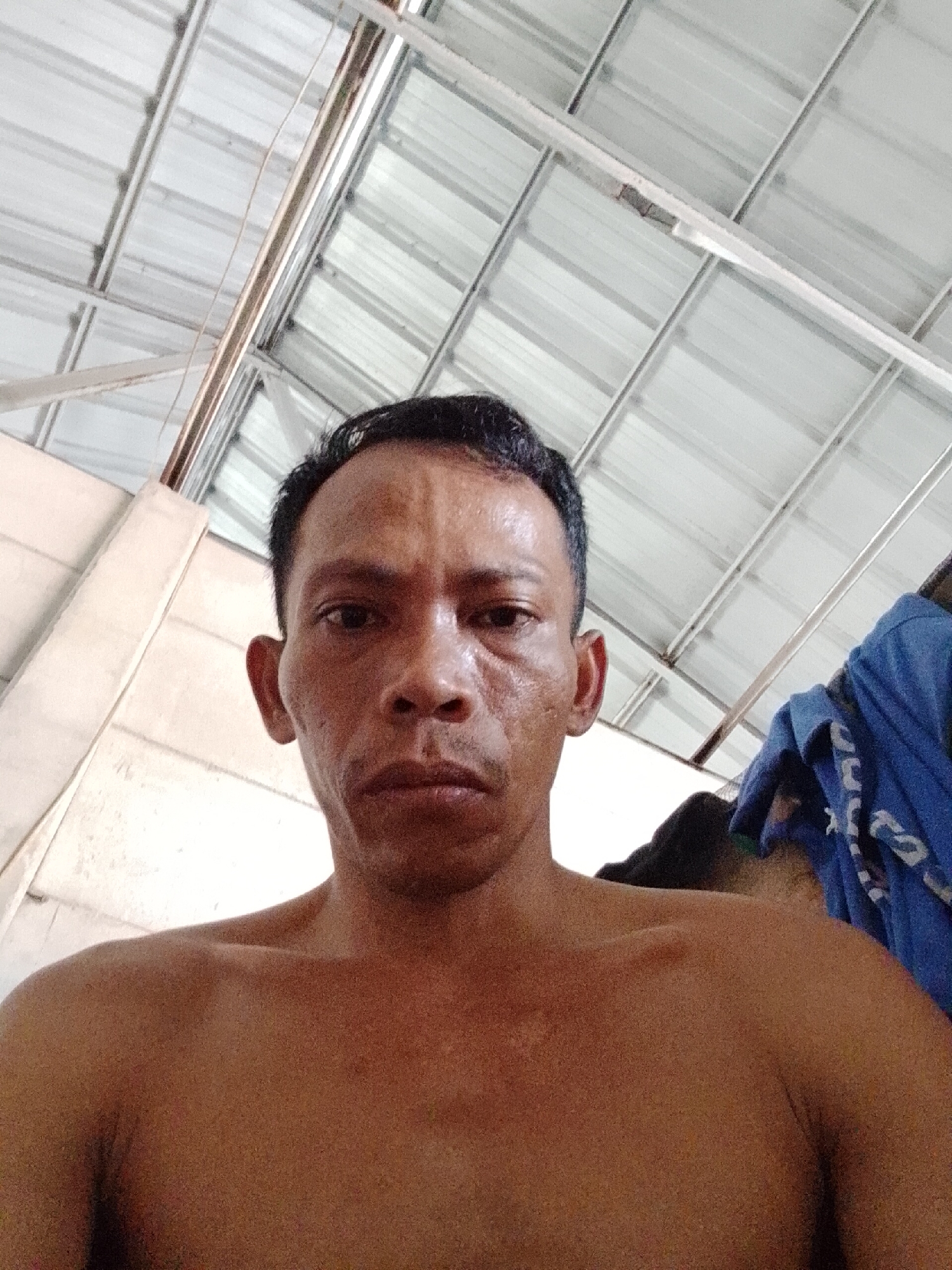 A shirtless man is photographed from a low angle, looking directly at the camera. His skin is tan, and he appears to be middle-aged. The background shows an indoor space with a corrugated metal roof, likely in the city of Bukit Ketok, Indonesia. A white wall is visible to the left, and a blue garment hangs on the right.