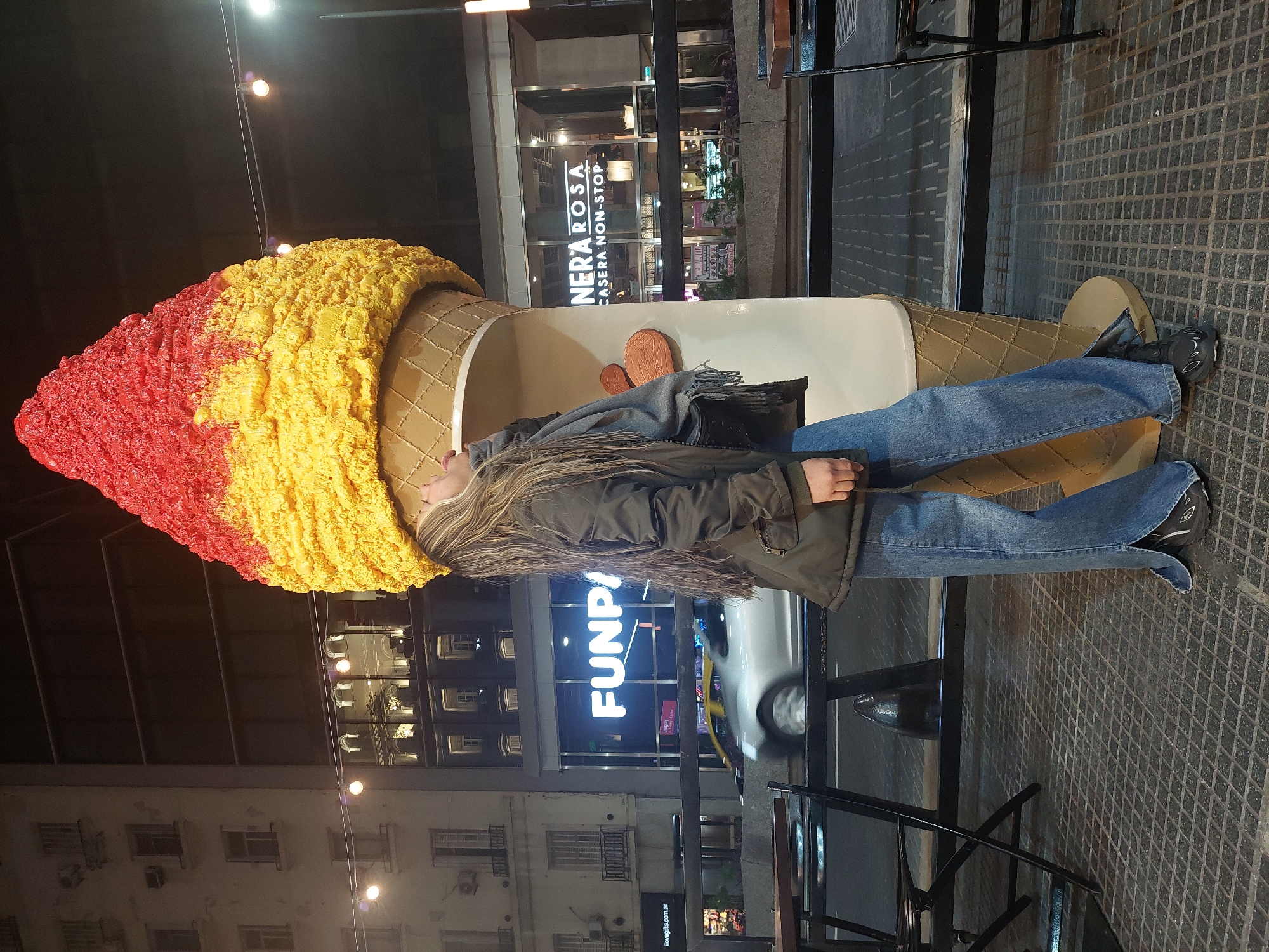 This vertical image captures an urban street scene at night in Buenos Aires, Argentina. The main subject is a person, appearing to be a young woman, standing next to a very large, oversized ice cream cone sculpture.
The person has long, light brown hair with highlights, wearing a dark olive-green jacket, blue jeans, and dark athletic shoes. A grey scarf is draped around her neck. She is looking upwards, towards the "scoops" of the ice cream cone.
The prominent ice cream sculpture features a textured red top scoop, blending into a yellow bottom scoop. The cone itself is a light brown, patterned with a waffle design. It appears to be an outdoor decoration or advertisement.
In the background, illuminated buildings line the street. One building clearly displays the text "NERA ROSA CASERA NON-STOP," suggesting a business specializing in homemade goods. Another building features a large, glowing sign that reads "FUNPA." Below, a smaller sign indicates "ilovegifts.com.ar," which confirms the Argentine location.
The street is dark, with artificial lights from the buildings and streetlights illuminating the scene. A blurred white car and a yellow vehicle (likely a taxi) are visible in motion on the street, indicating active city life. The ground is paved with dark, square-patterned tiles. Dark metal structures, possibly railings or outdoor seating, are visible to the right. The person's jacket and scarf suggest the weather might be cool.