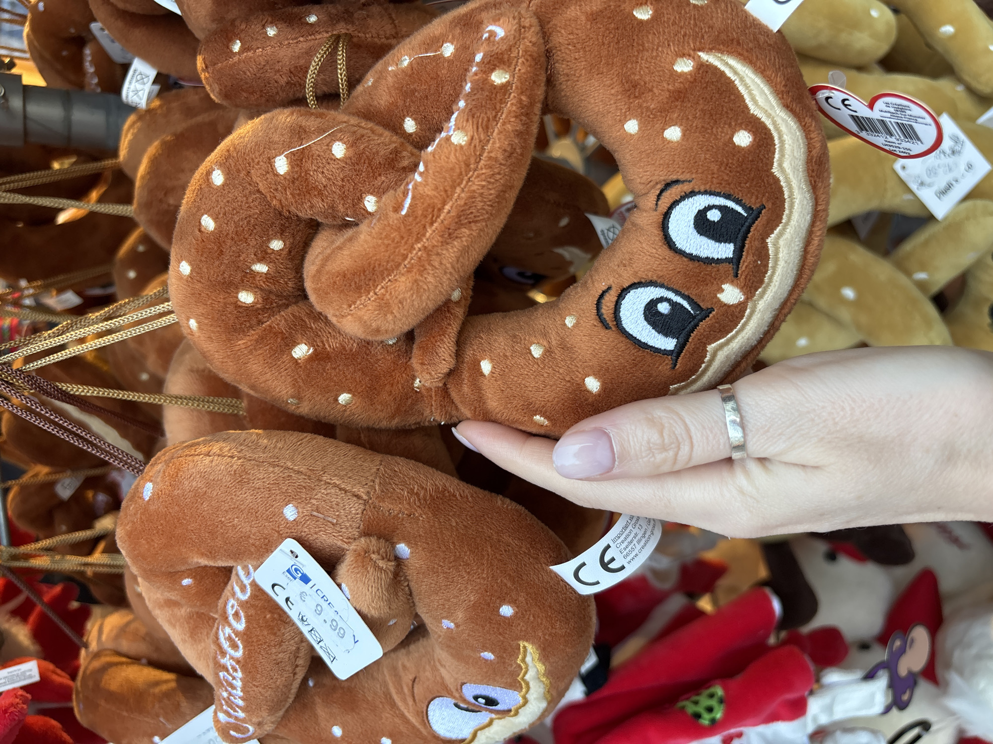 A close-up shot shows a person's hand holding a plush pretzel toy with embroidered white dots and a smiling face. The pretzel is a warm brown color and appears to be a Christmas ornament. It is hanging amongst other similar plush pretzels. A price tag is visible on one of the pretzels, reading "6.99" and "CE". In the background, other plush toys, including red and white characters, are visible. The scene is likely a Christmas market in Strasbourg, France, given the characteristic style of the plush toys and the presence of the "CE" marking.