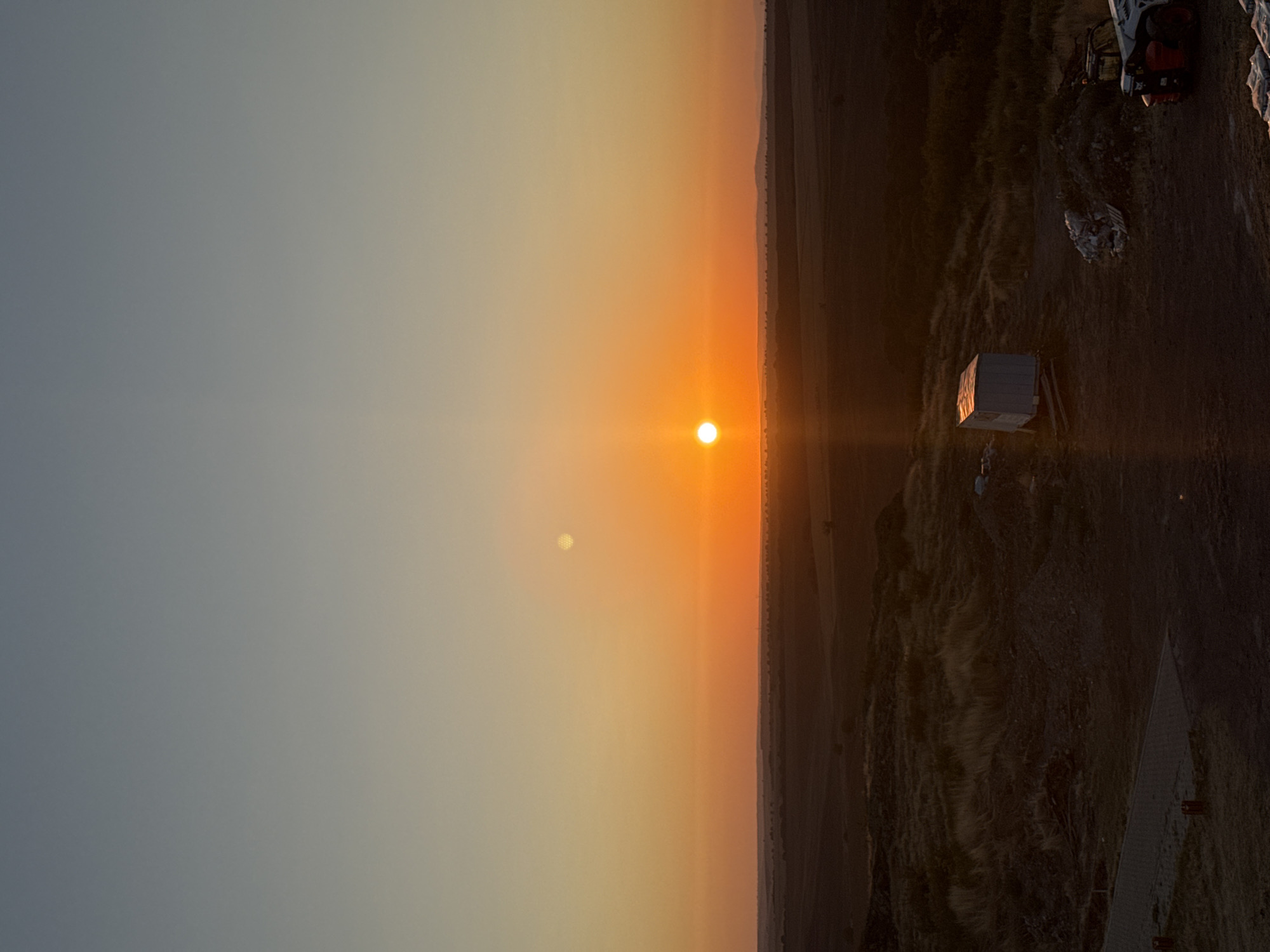 The image depicts a sunset over a rural landscape, presumably near the Bulgarian city of Rusokastro. The sky displays a gradient of colors, transitioning from a pale blue at the top to vibrant orange near the horizon. The sun, a bright orange orb, is low in the sky, casting a warm glow. A faint, circular artifact is visible in the upper right portion of the sky, possibly due to lens flare or a celestial object.
The foreground features uneven, dry terrain with patches of grass and scattered debris. A small, white portable structure, resembling a shed or temporary shelter, sits on a wooden base. Adjacent to it are piles of dirt and rubble. In the lower right corner, a portion of a vehicle, possibly a construction vehicle or utility vehicle, is visible. The overall environment suggests an undeveloped or construction area. The landscape stretches out to the horizon, with subtle undulations and distant, low-lying hills. The lighting indicates either dawn or dusk.