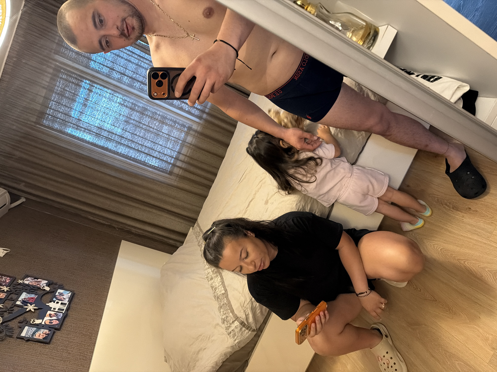 A man in boxers, a child in a pink dress, and a woman in a black shirt are in a bedroom. The man is holding a smartphone and looking into a mirror. The child is lying on a bed, and the woman is sitting on the floor, also looking at a smartphone. There are several framed photos on the floor next to the woman. The room appears to be well-lit, suggesting it is daytime. The overall atmosphere seems casual and domestic. The text "POLO ASSN." is visible on the man's boxers.