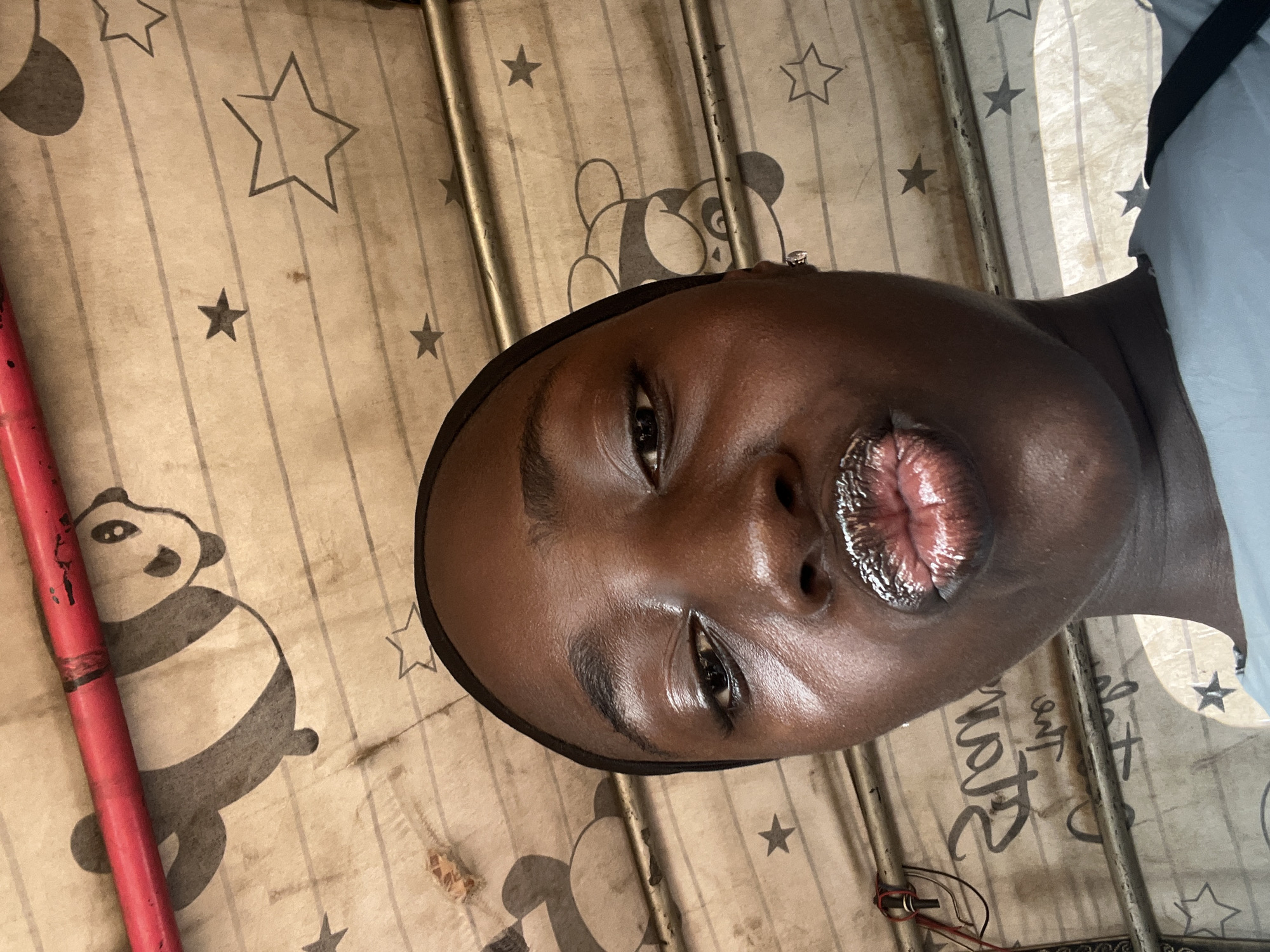 The image is a close-up, low-angle selfie of a young Black woman in Galadimawa Village, Nigeria. She is wearing a black hijab and a light blue shirt. Her lips are pursed and appear to be covered in glitter or a shimmery lip gloss. She is wearing small stud earrings. The background is a fabric material with a pattern of pandas and stars, and some horizontal lines that suggest ruled paper. A red rod is visible at the top of the image, and metal bars run horizontally across the frame. Some wires are also visible on the left side. The lighting is bright and appears to be natural, possibly from sunlight.