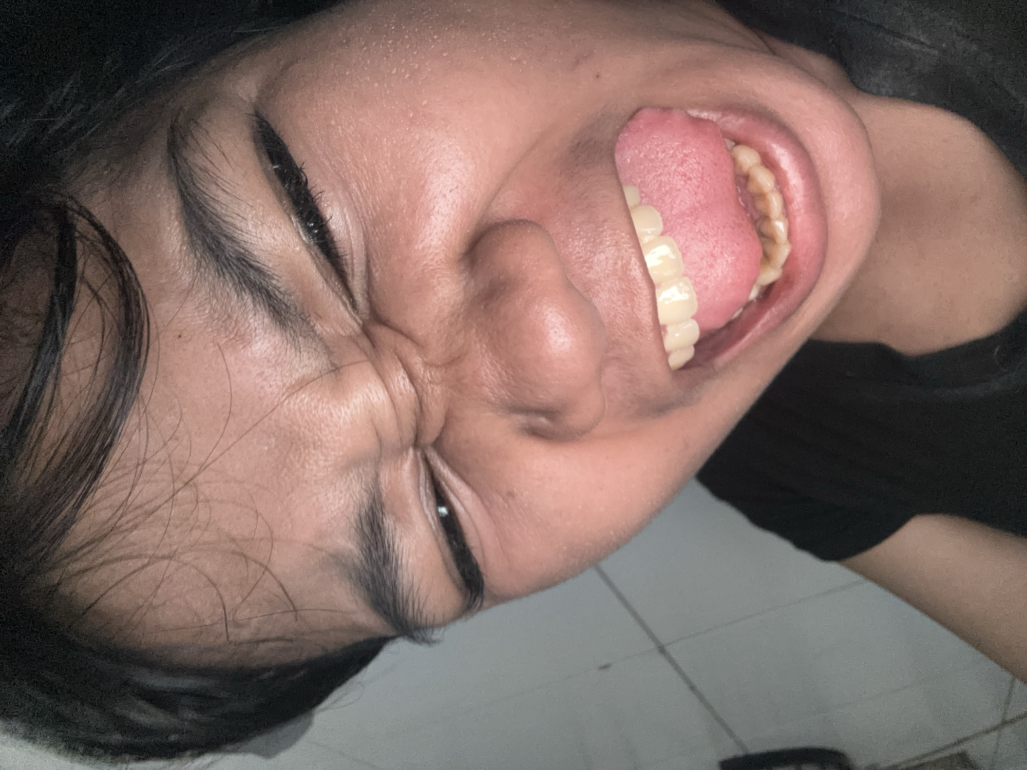 A close-up, low-angle shot shows the face of a person lying down with their mouth open and tongue extended. The person's eyes are squinted, and their teeth are visible. The background is a tiled floor. The image appears to have been taken indoors in a residential setting in North Jakarta, Indonesia.