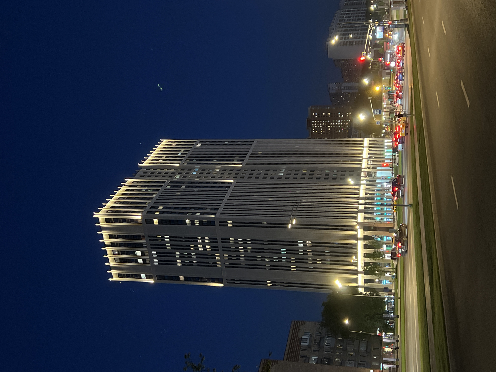 A tall, modern building with horizontal lines and illuminated windows stands beside a wide, multi-lane road in an urban setting at night. The building's lights contrast with the dark blue sky. The road is lined with trees and streetlights and has traffic. Other buildings are visible in the background.