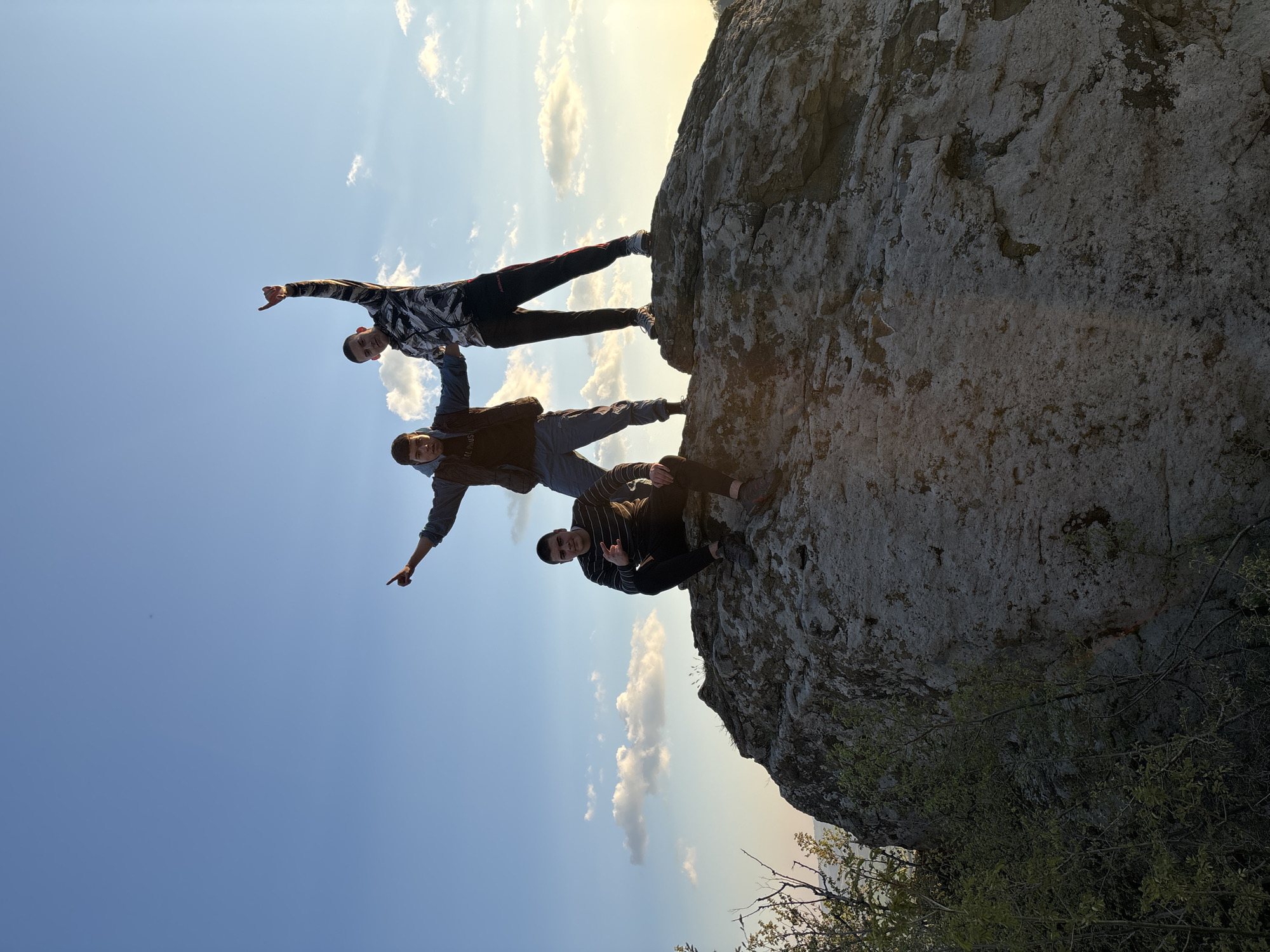 Three young men are posing on a large rock formation. The man at the top stands with his arms outstretched, pointing in opposite directions, and appears to be balanced precariously. The man in the middle mirrors this pose, also pointing in opposite directions. The man at the bottom is seated and making a hand gesture. The sky is blue with scattered white clouds. The background suggests a natural environment. Based on the prompt, the location is Goritsa, Bulgaria.