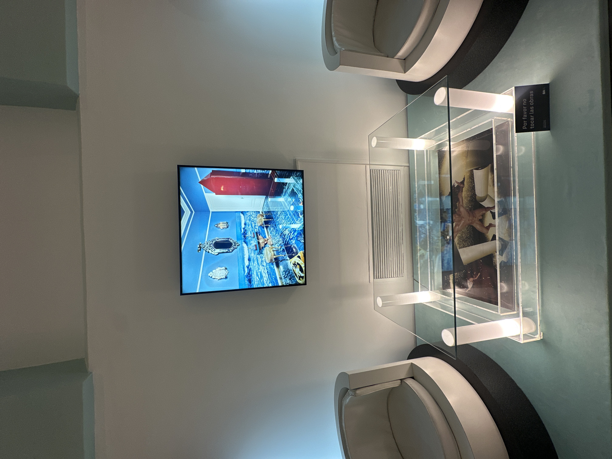 Indoor scene showcasing an art display. A flat-screen TV mounted on a white wall displays an image of an interior with a red-ceilinged room and ornate furniture, potentially an artistic creation. Beside the TV is a ventilation unit or decorative panel. The display includes a clear, box-shaped showcase with white cylindrical supports, filled with printed materials or artwork. A small sign is attached to the showcase, bearing the Spanish text "Por favor no tocar las obras" (Please do not touch the works). Two white, semi-circular chairs with black bases flank the showcase. The color scheme is predominantly white, light blue, and gray. The location seems to be a museum or art gallery in Buenos Aires, Argentina. The display is focused on art and interactive viewing via the television screen. No people are directly visible in the frame.