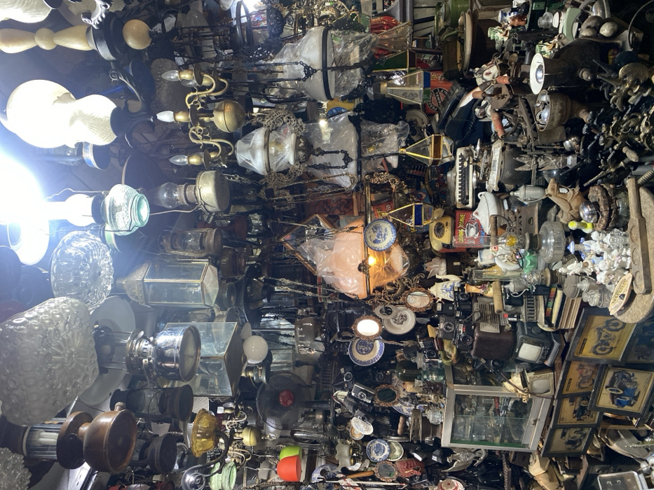 The image shows an interior scene of a cluttered shop in Surakarta, Indonesia, located at coordinates -7.569 latitude and 110.822 longitude. The shop is filled with an array of antique oil lamps, chandeliers, and various other trinkets, including cameras, framed photographs, and other miscellaneous objects. The ceiling is covered in a dense network of electrical wires and hanging fixtures. The shop is dimly lit and the atmosphere is somewhat chaotic.