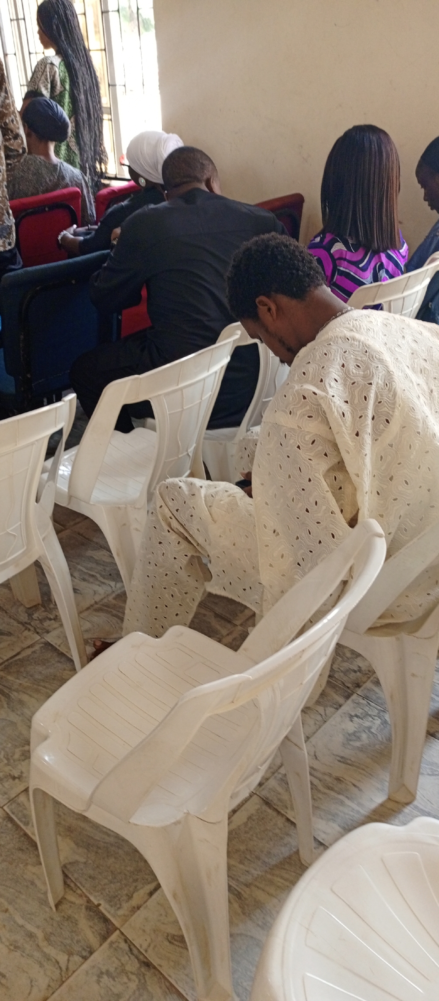 Location: Keffi, Nigeria.
Scene: A gathering of people indoors. The scene includes several individuals seated on white plastic chairs.
People: Multiple individuals are present. A man in the foreground wears an off-white patterned outfit, possibly traditional attire. Other individuals are visible in the background, some wearing black clothing, white head coverings, and patterned clothing.
Objects: White plastic chairs are scattered throughout the room. Some of the individuals are seated on red chairs.
Environment: The room appears to be brightly lit, with light-colored walls. The floor has a tiled pattern. The area suggests a community center or meeting space.