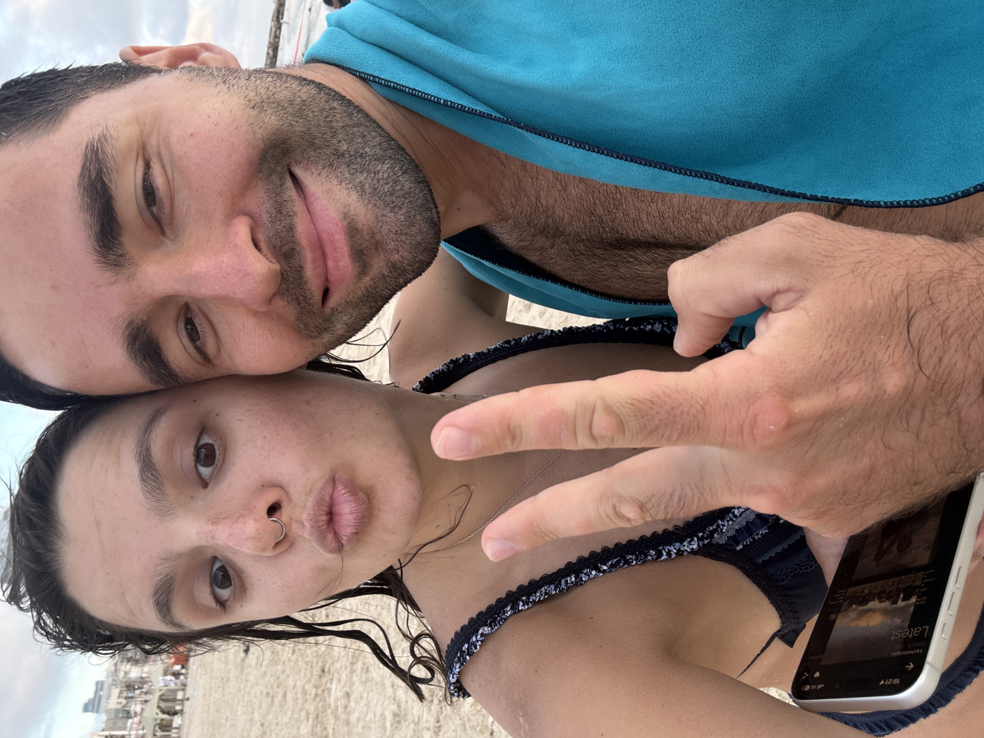 This media file is a selfie taken by two people on a beach, likely in Tel Aviv, Israel, given the geotag. The image is oriented vertically, with the subjects' faces filling the majority of the frame.
The person in the foreground on the right is wearing a dark bikini top with a patterned trim. Their hand is raised, and they are making a peace sign with two fingers. Their lips are pursed in a "duck face" pose. They have a nose ring and their hair is dark and appears wet.
The second person, positioned behind and to the left, is also looking towards the camera. They have dark hair and a beard, and are wearing a bright blue towel draped over their shoulders. Their eyes are open, and they have a slight smile.
The background shows a sandy beach with some faint structures and possibly people in the distance, suggesting an outdoor, daytime setting. The sky is overcast, indicating possibly cloudy weather. The time stamp on the phone visible in the lower right corner reads "19:21," suggesting it is late afternoon or early evening. The phone screen displays the word "Latest," possibly referring to a news feed or social media content. The overall mood appears casual and relaxed, consistent with a beach outing.