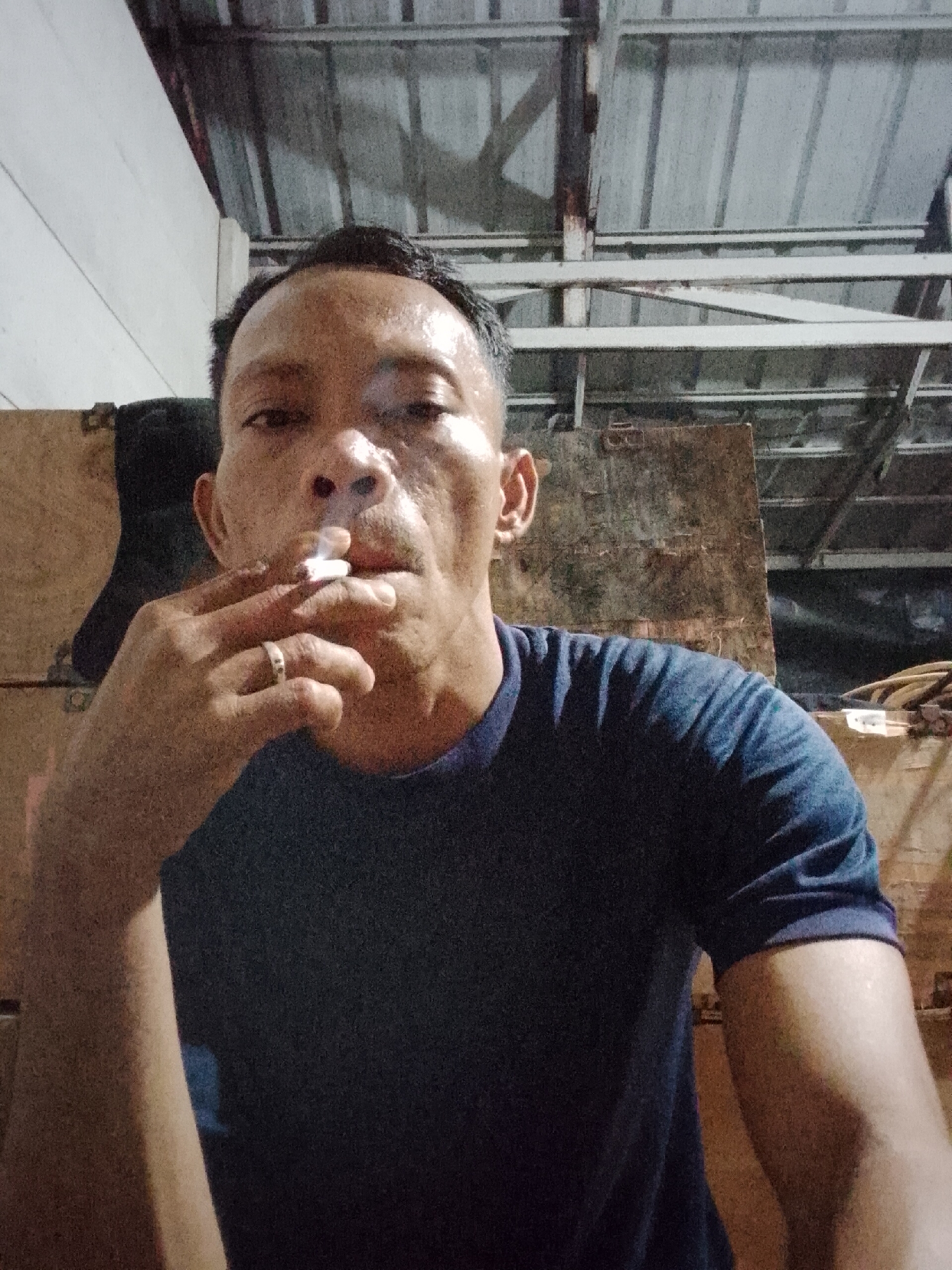 A close-up, over-the-shoulder selfie shows a middle-aged Indonesian man smoking a cigarette indoors. He is wearing a dark blue t-shirt and a ring on his left hand. The background appears to be a dimly lit storage area with wooden crates and exposed metal roofing. The man's face is turned towards the camera, and he is exhaling smoke. The image does not provide information about the specific location within Bukit Ketok, Indonesia.